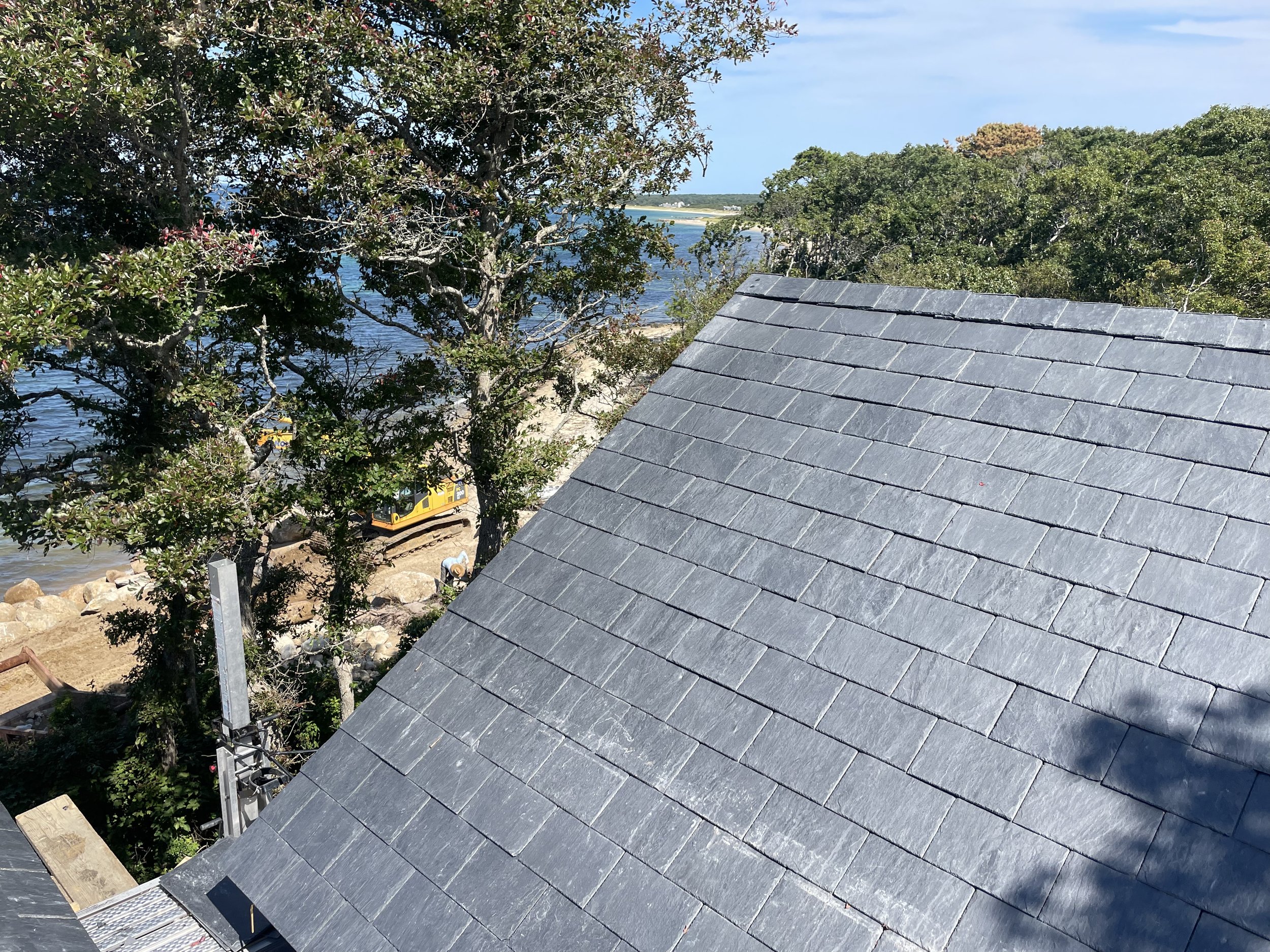 View of a rooftop with gray shingles, trees, water, and distant shoreline with buildings, seen from a higher vantage point.