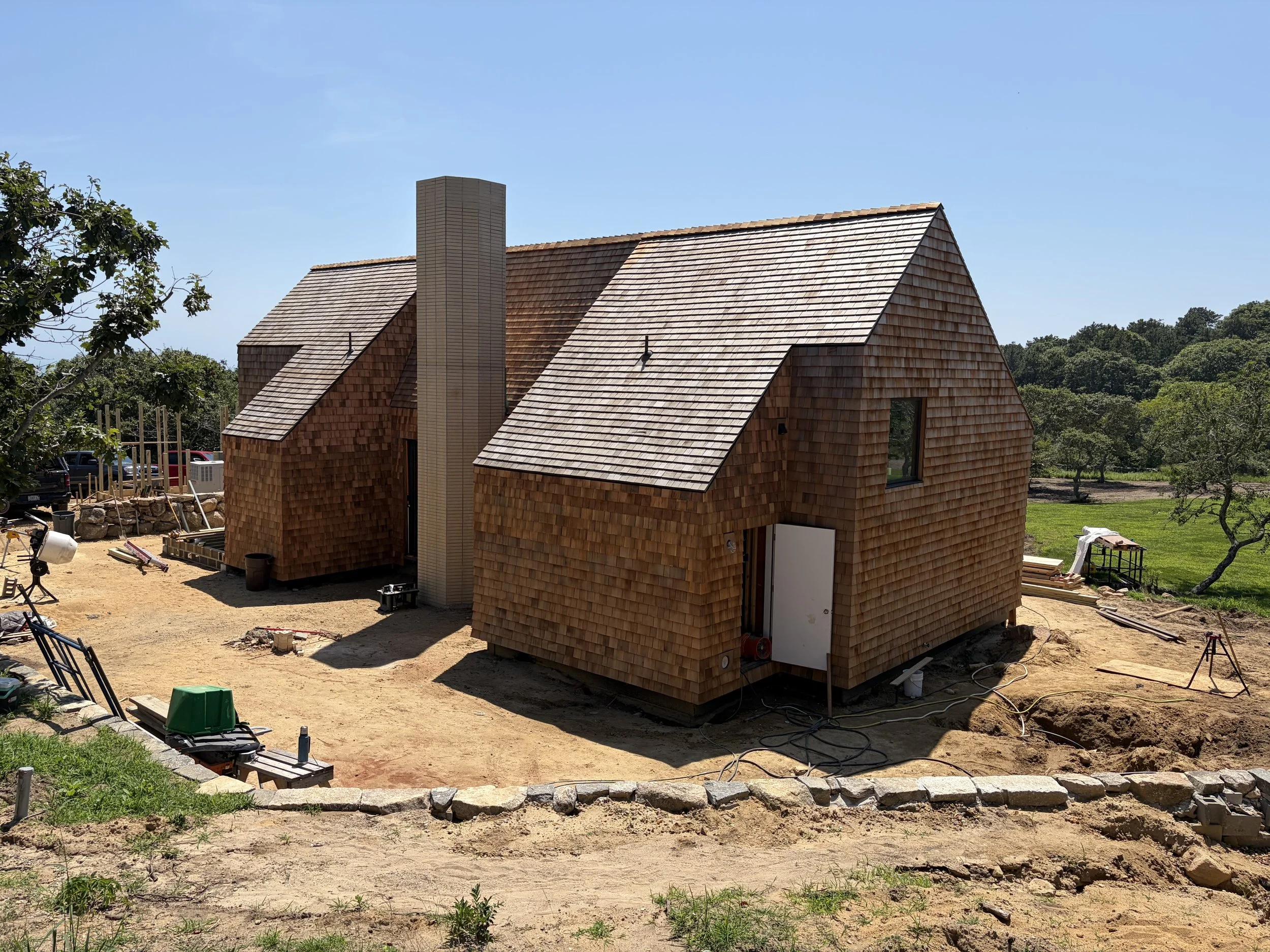 A two-story house under construction with wooden shingle siding and a steep gable roof. The house features a brick chimney and windows, with construction tools and materials surrounding the site. The background includes trees and a clear blue sky.