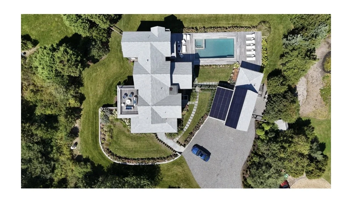 Aerial view of a house with a backyard featuring a swimming pool, patio, and solar panels on the roof, surrounded by greenery and trees.
