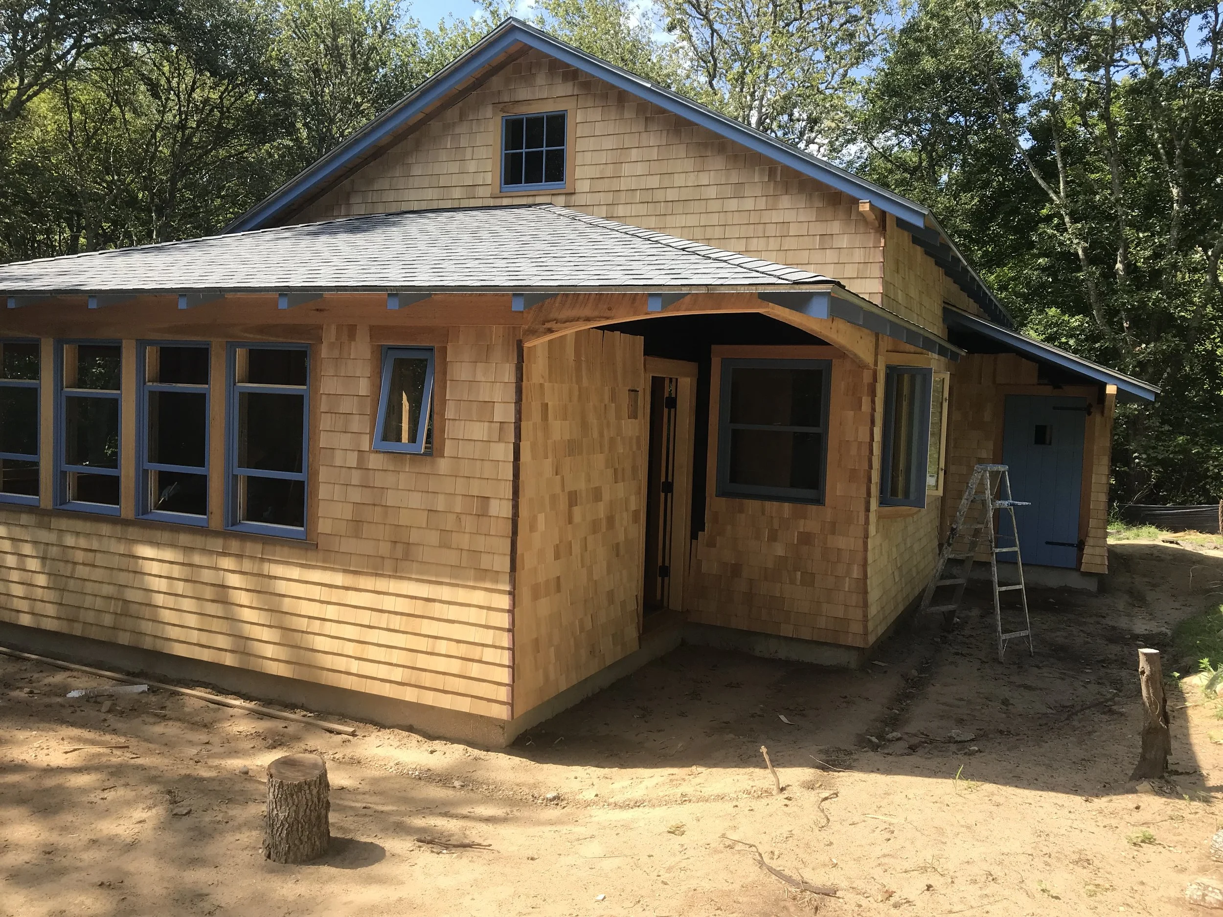 A house under construction with wooden siding and multiple windows. The building is surrounded by trees and construction equipment, including a ladder.
