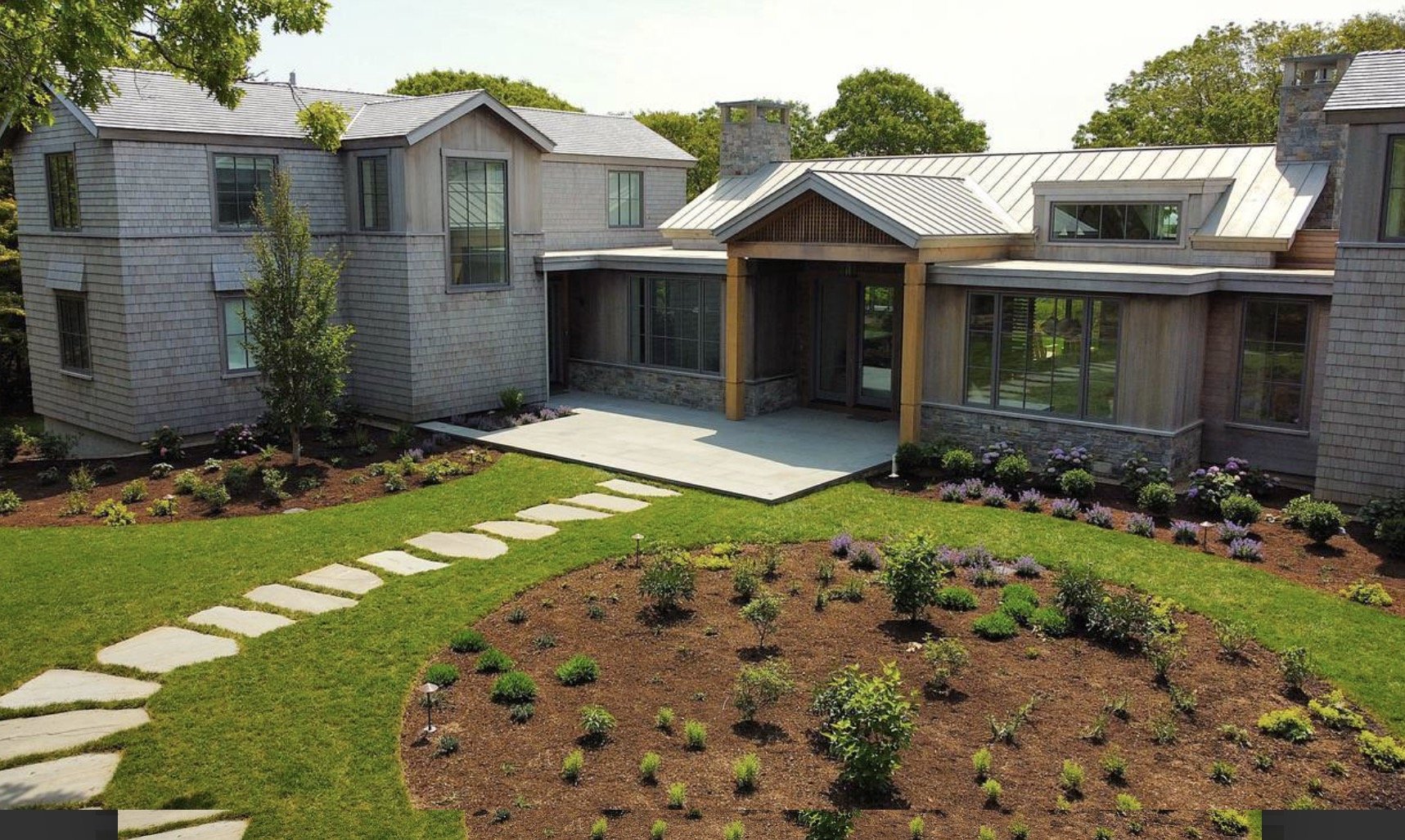 A modern two-story house with gray siding, large windows, a metal roof, and a chimney, surrounded by a well-maintained garden with a stone walkway.