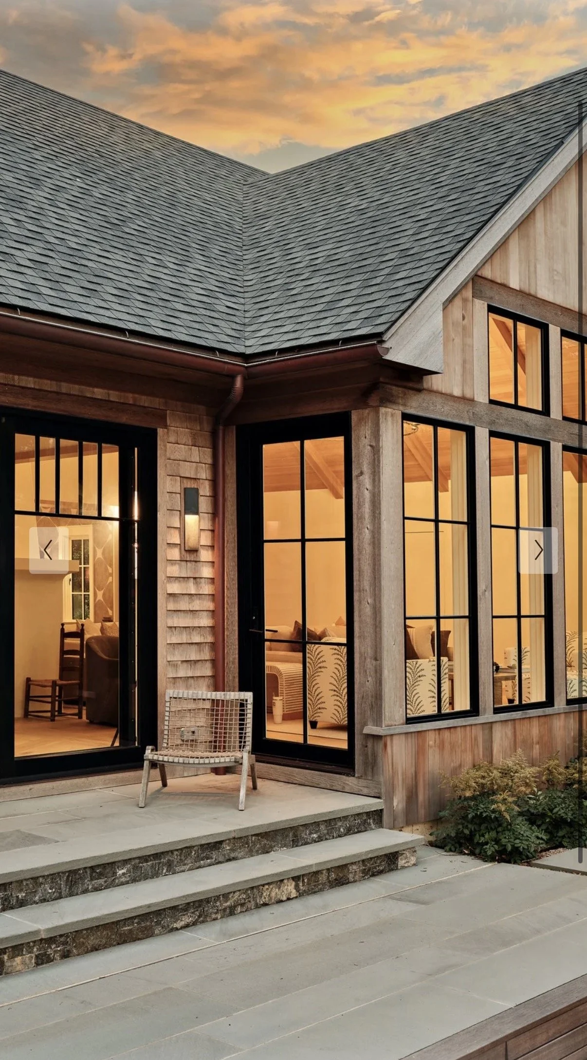 Close-up of a modern house with large glass windows, wooden exterior, and a sharply pitched roof, at sunset.