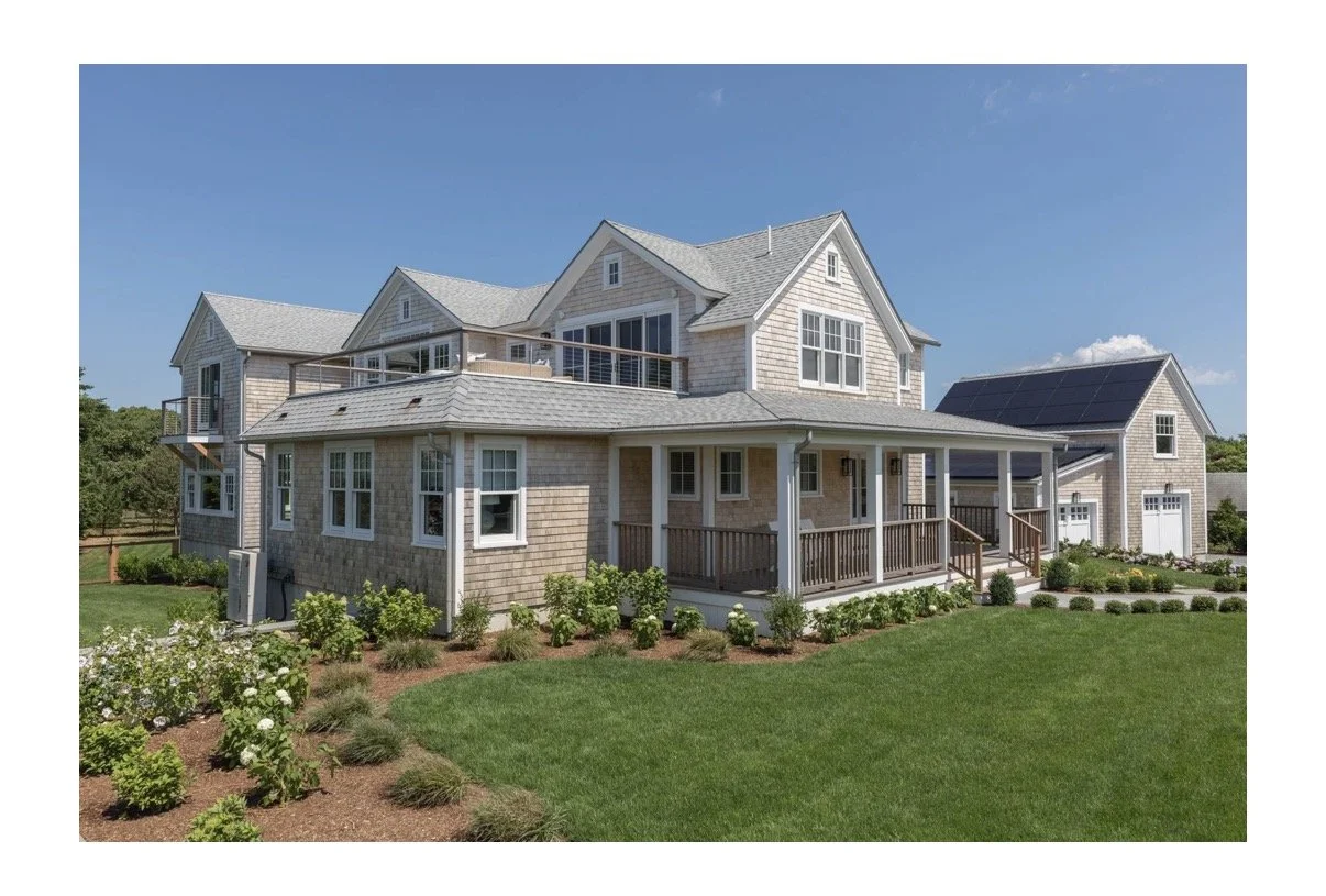 A large, modern house with gray shingles, multiple gabled roofs, a wrap-around porch, and solar panels on the roof in a well-manicured yard.