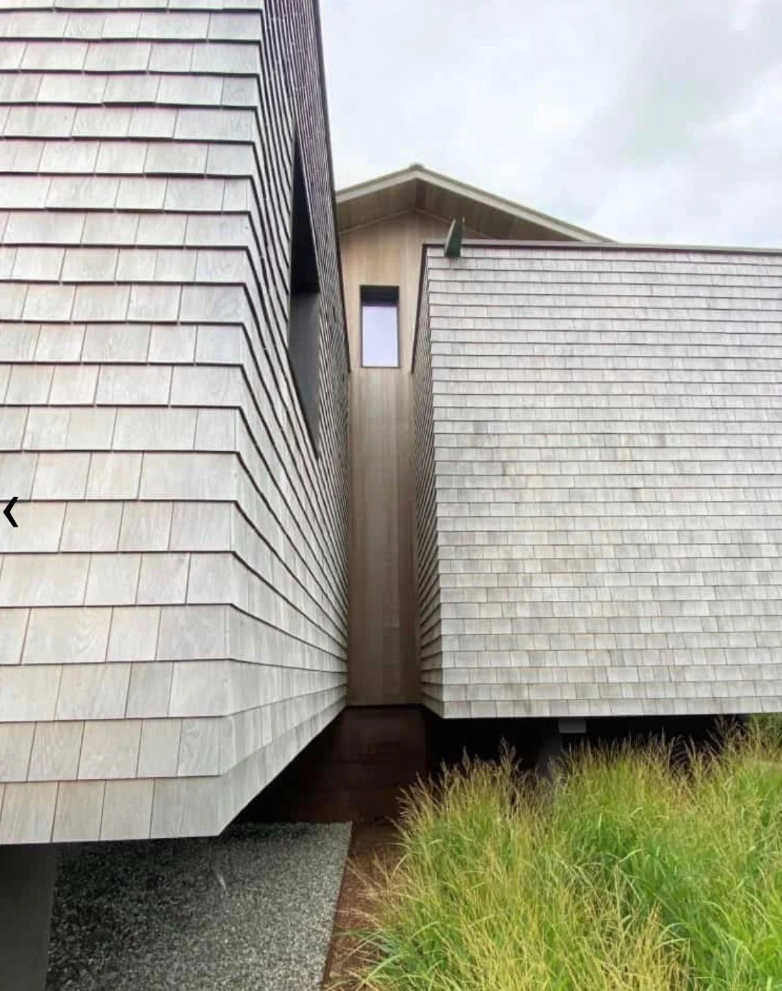 Two modern houses with gray shingle siding facing each other, with a narrow gap between them leading to a small protruding section of house in the background, set against an overcast sky with green grass at the base.