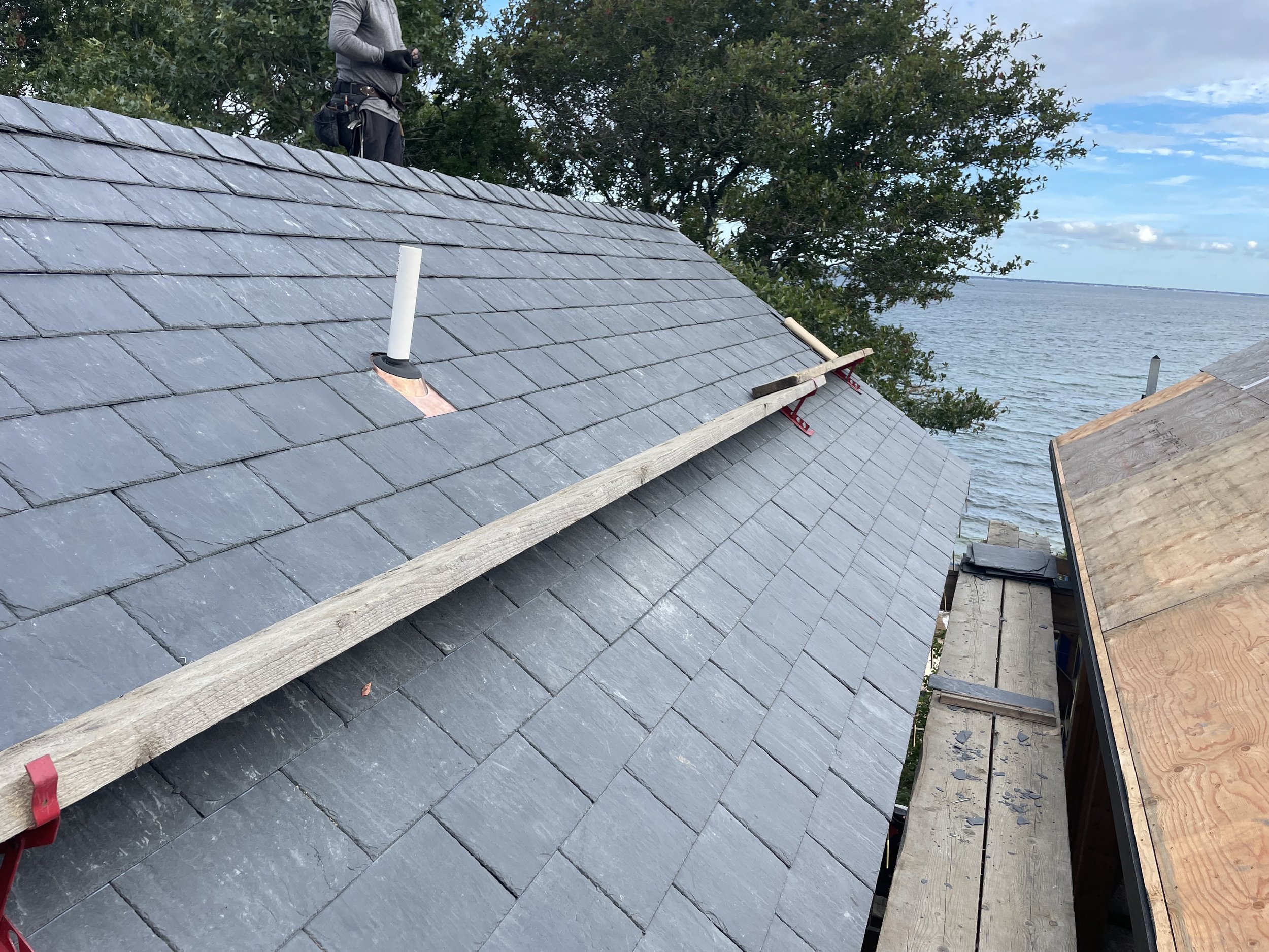Person working on installing slate roof tiles near a body of water, with a lake and tree in the background.
