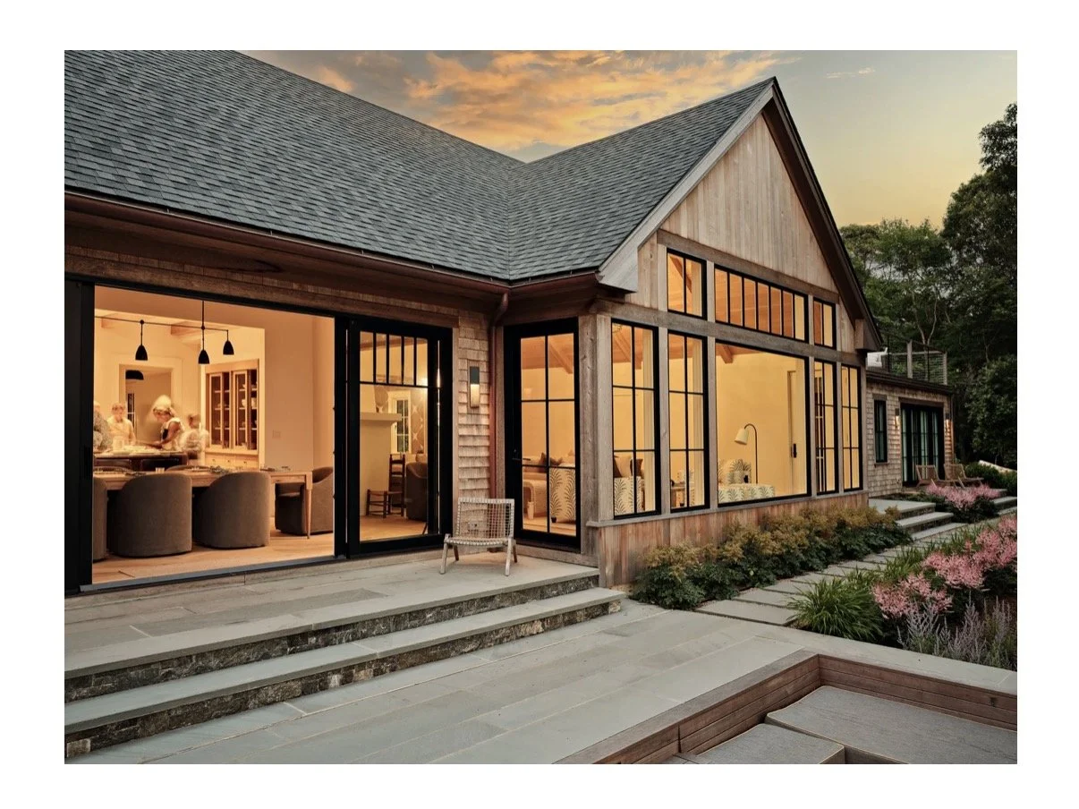 Modern house with large windows at sunset, interior visible with dining and living areas, surrounded by landscaped garden with flowers and bushes.