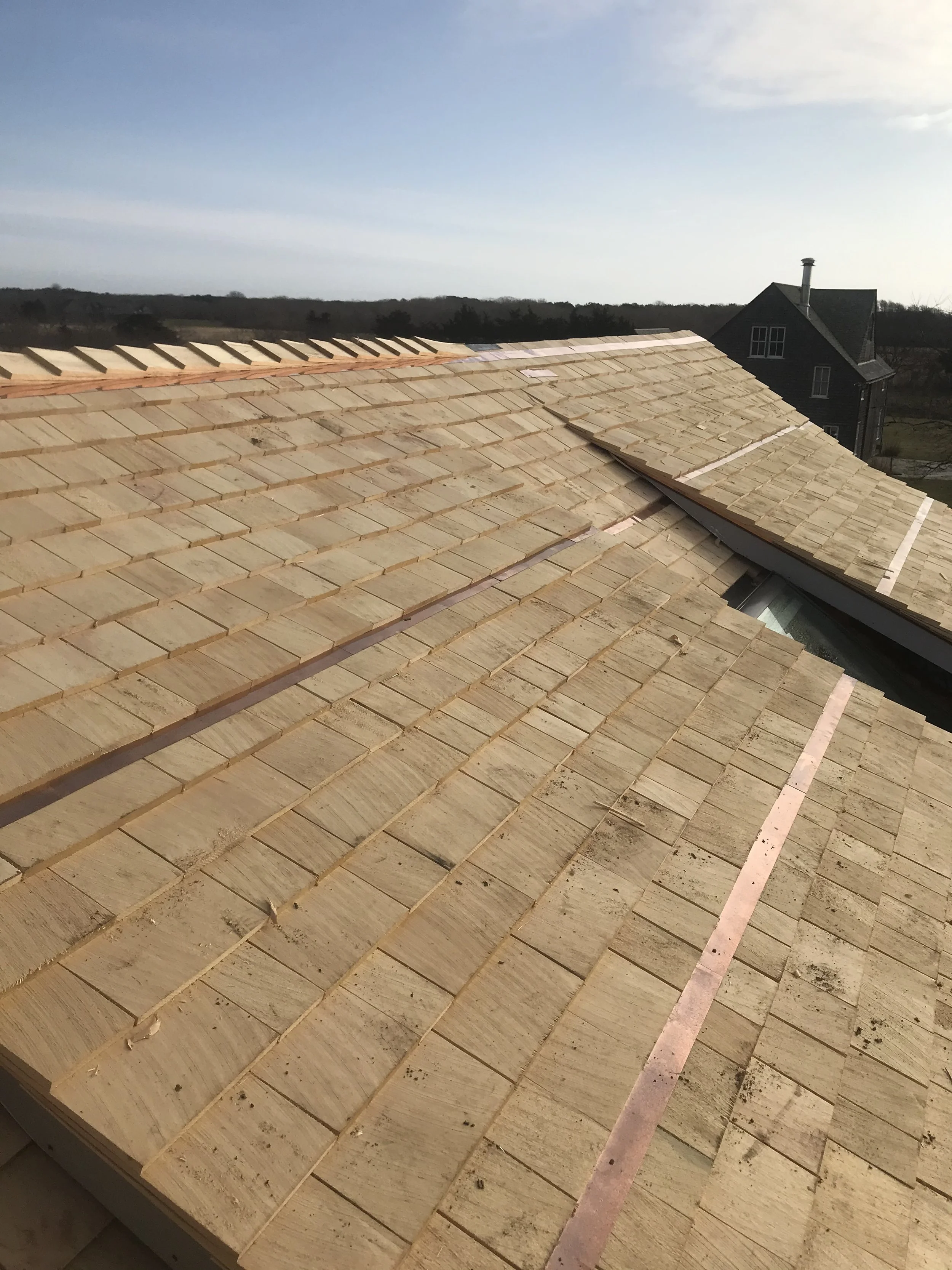 View of a house roof under construction with unfinished wooden shingles installed, overlooking a rural landscape and a neighboring house.