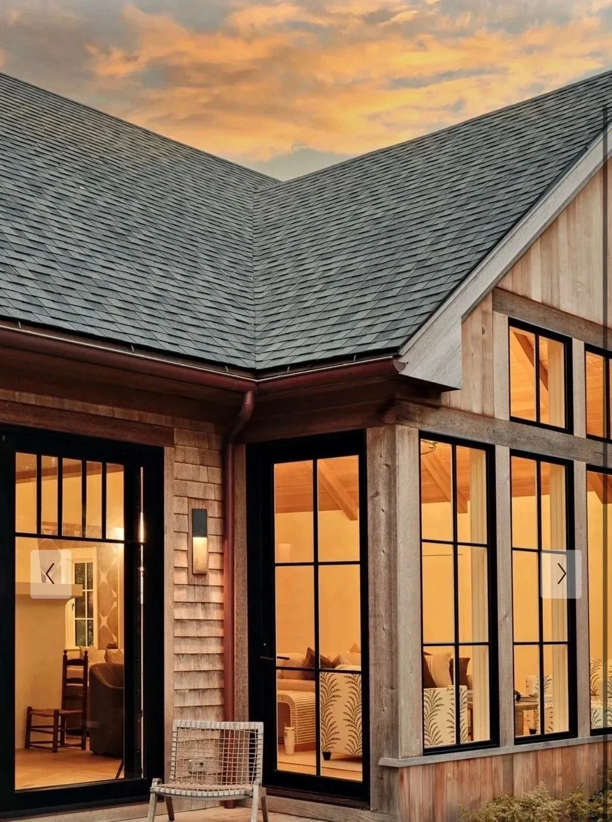 Close-up of a modern house with large black-framed windows, wooden siding, and a slate roof under a sunset sky.
