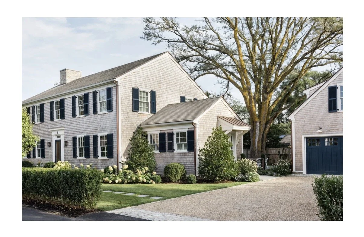 A large house with light-colored siding, multiple windows with black shutters, a driveway, and a large tree in a landscaped front yard.