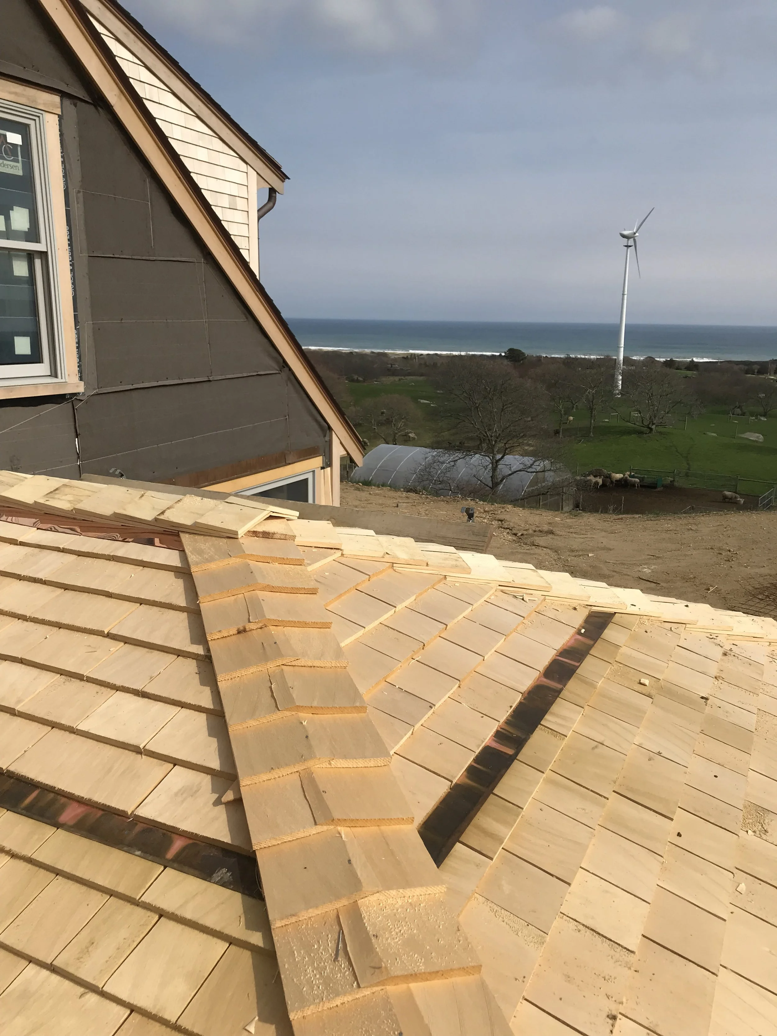 Roof shingles stacked on a construction site with a view of a green field, wind turbine, and ocean in the background.