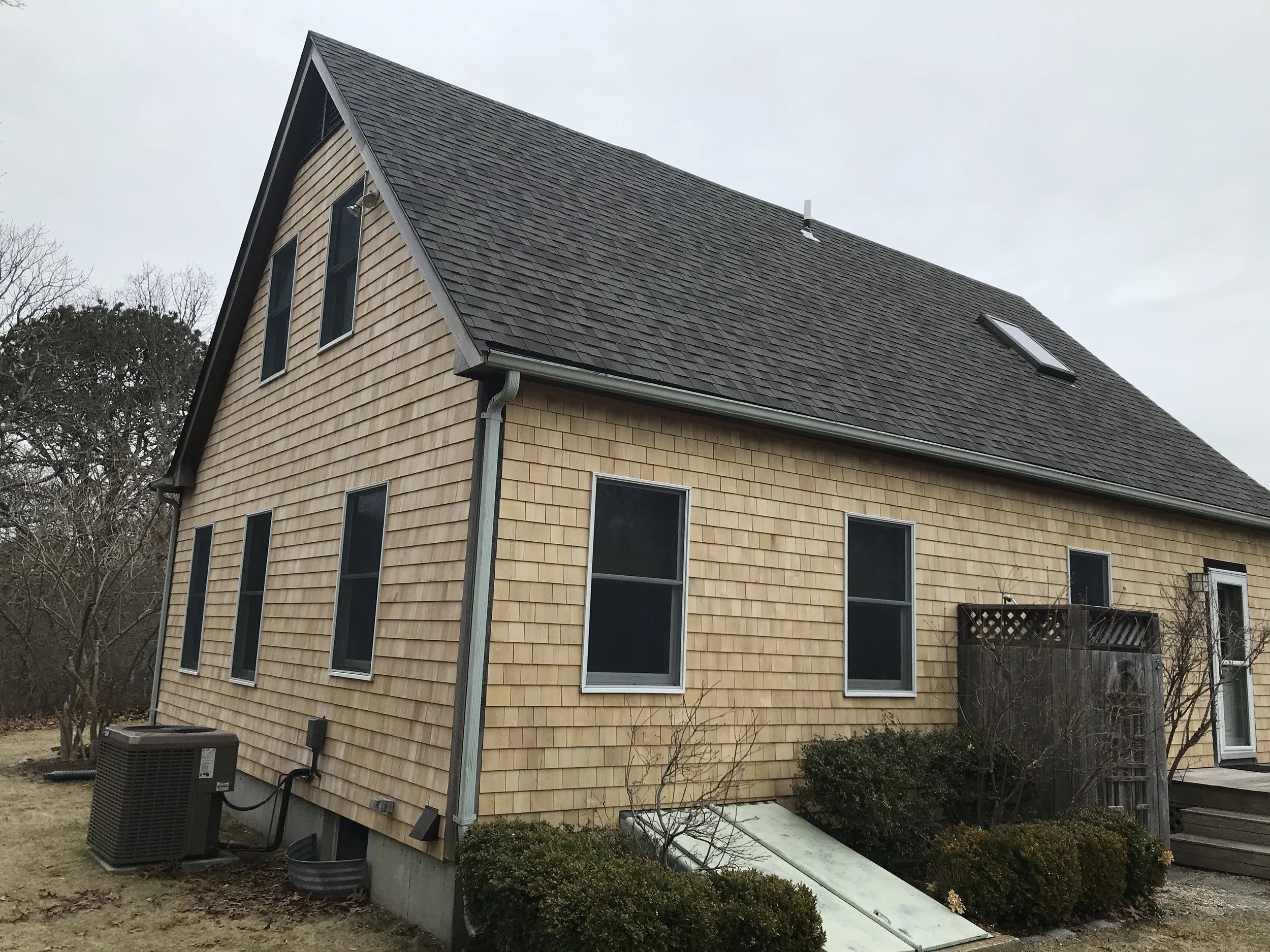 Side view of a house with beige wood shingles, multiple windows, and a steep, dark gray shingle roof with skylights and a vent, surrounded by leafless trees and bushes.