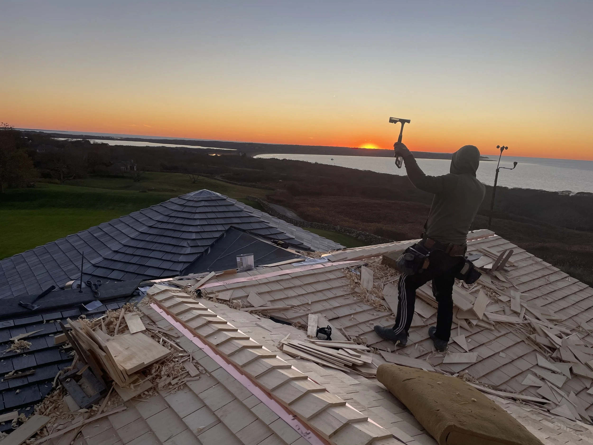 Person wearing a hoodie and striped pants working on a roof with shingles, holding a hammer, during sunset with a view of a large body of water and flat landscape in the background.