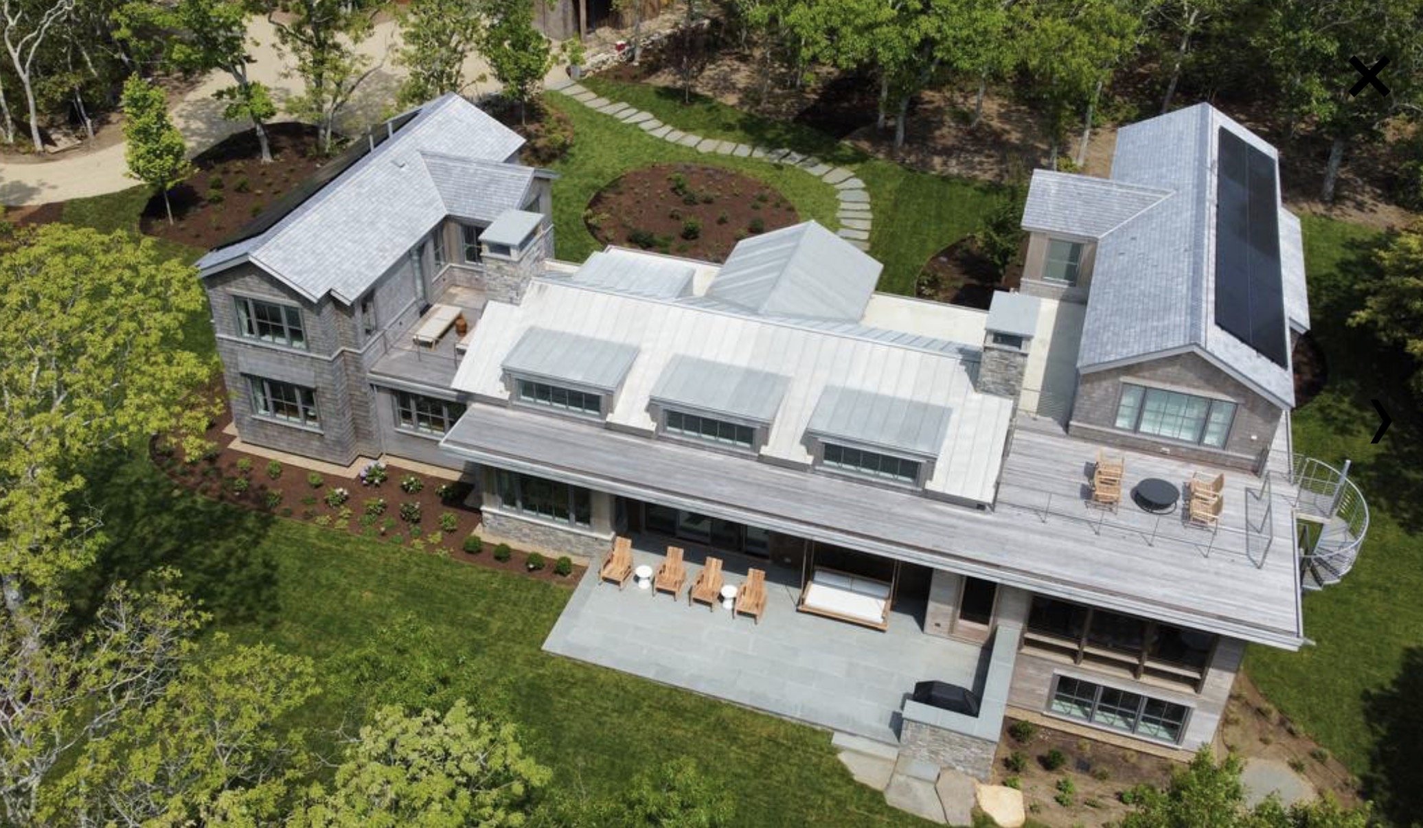 Aerial view of a large house with gray roof, surrounded by green trees and lawn, featuring a patio with outdoor furniture and a landscaped yard with a stone pathway.