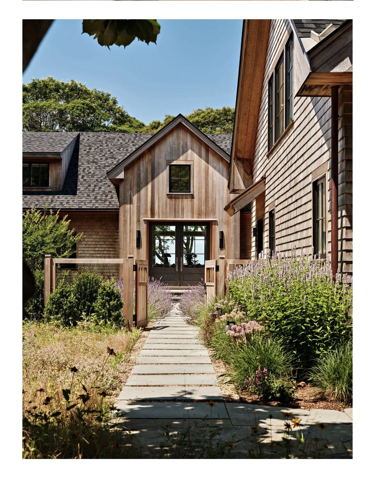 A wooden house with a modern design, surrounded by landscaped garden with flowers and bushes, under a clear blue sky.