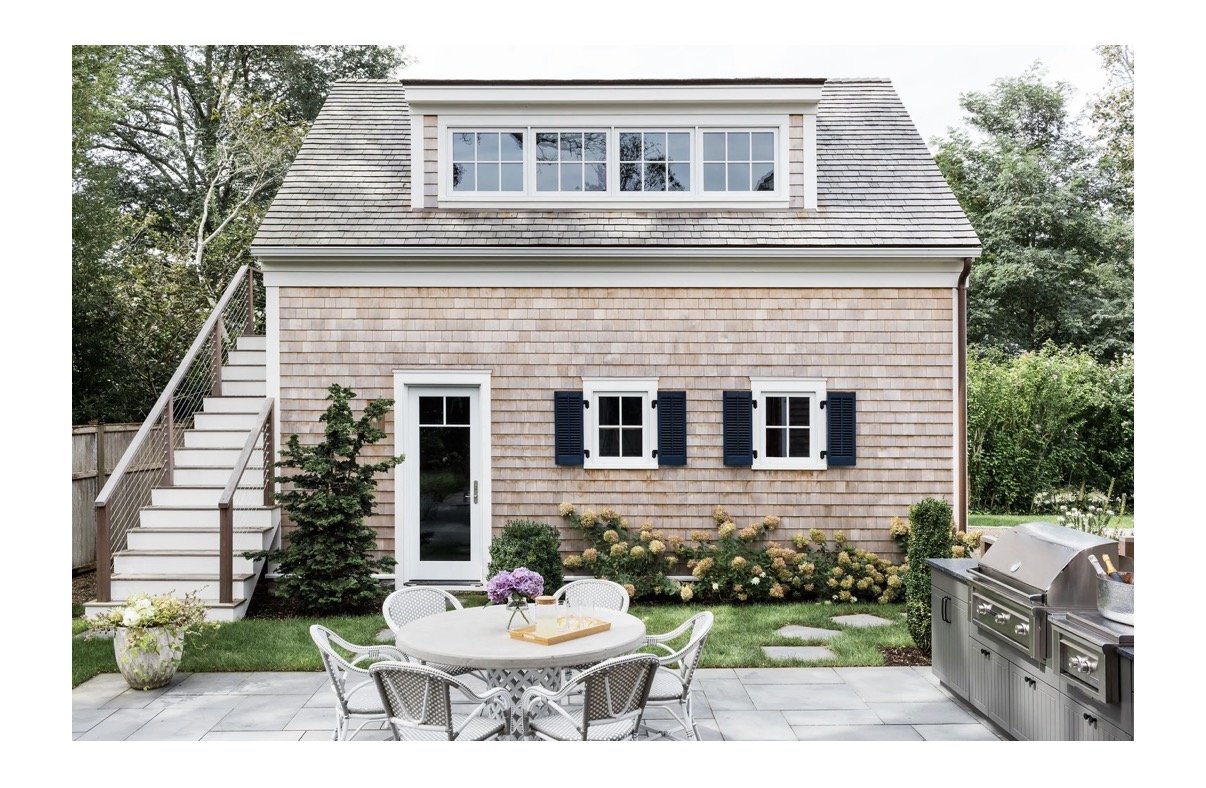Backyard of a house with a patio, outdoor furniture, and a grill. The house has light-colored shingles, three small windows with blue shutters, a glass door, and a large dormer window on the roof. There are trees and bushes surrounding the area.