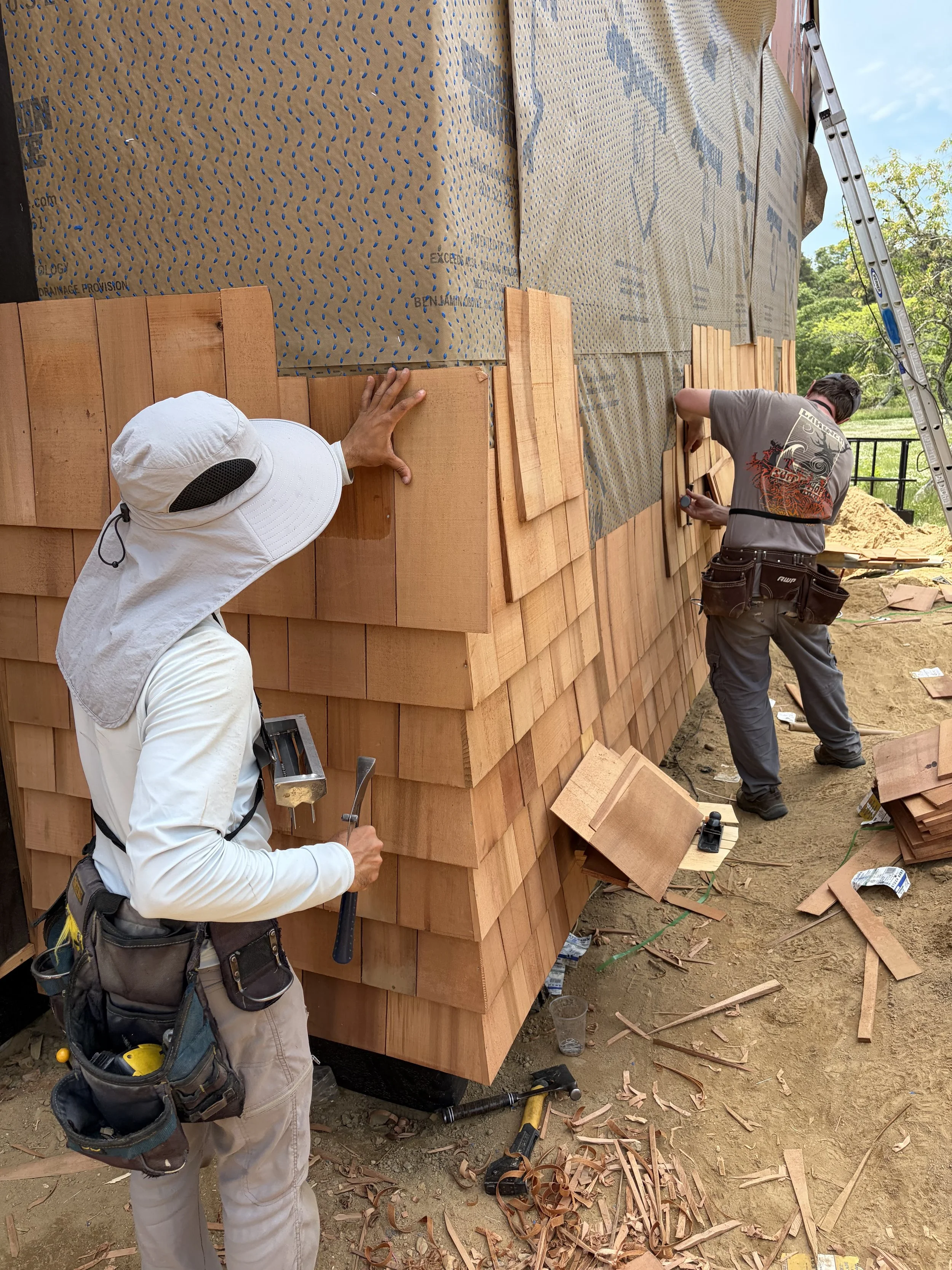 Two workers installing wooden shingles on the exterior wall of a building, with tools and wood debris on the ground.