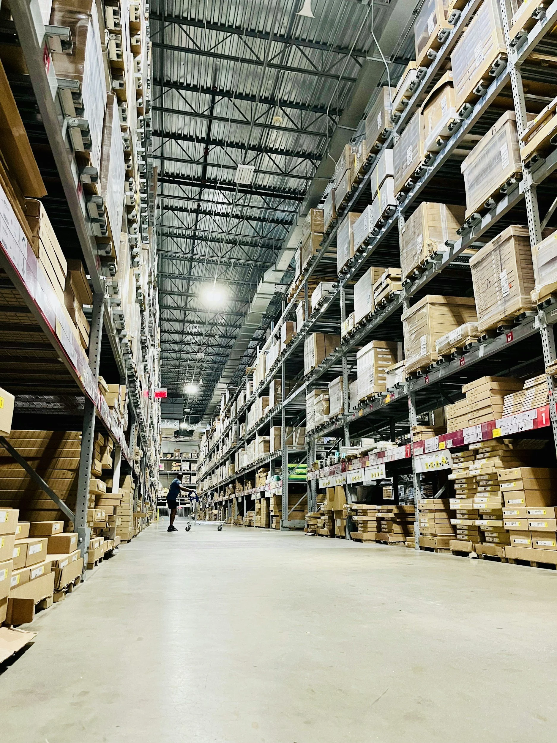 Employee walking through a large warehouse aisle with shelving and inventory.