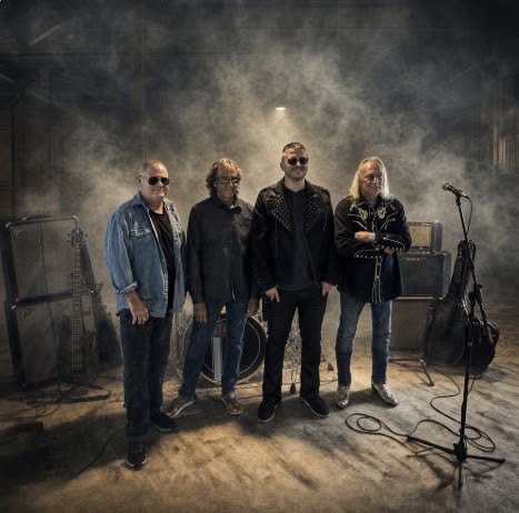 Four men standing together on a stage with musical equipment, in a smoky environment, posing for a photo.