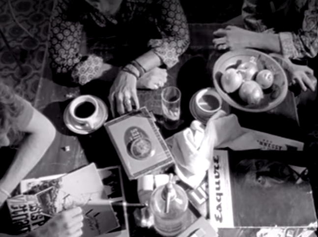 A top-down view of a table with people, a bowl of apples, magazines, a drink, and a napkin dispenser.