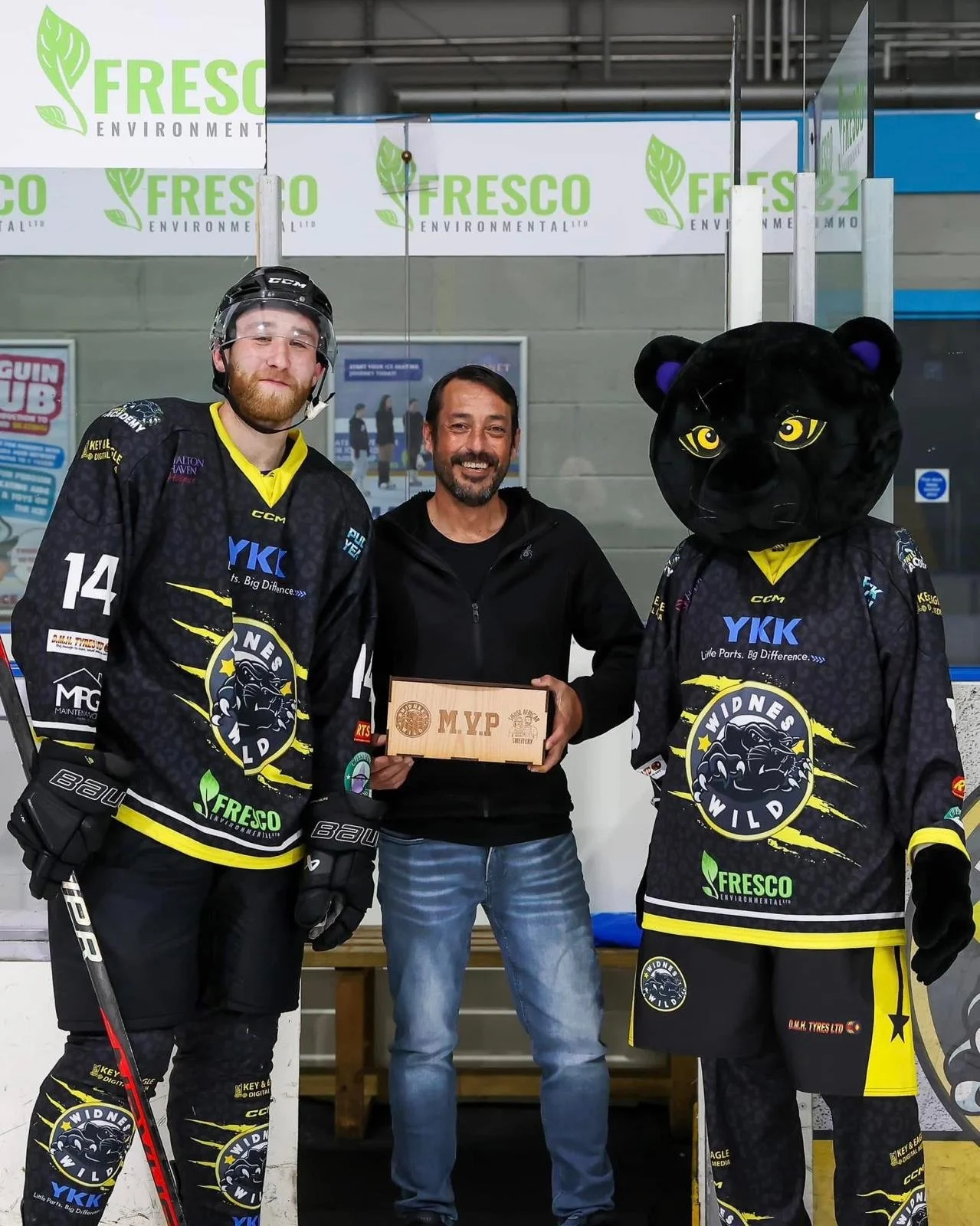 Two ice hockey players in black and yellow jerseys, one with a lion mask, stand alongside a man holding a wooden plaque that reads MVP, while posing with a man. They are in an indoor ice rink with a banner reading 'FRESCO ENVIRONMENTAL' in the background.