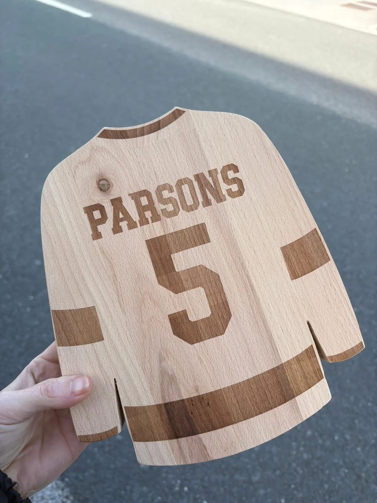 A wooden sign shaped like a football jersey with the name 'Parsons' and the number '5' engraved on it, held over a paved surface.