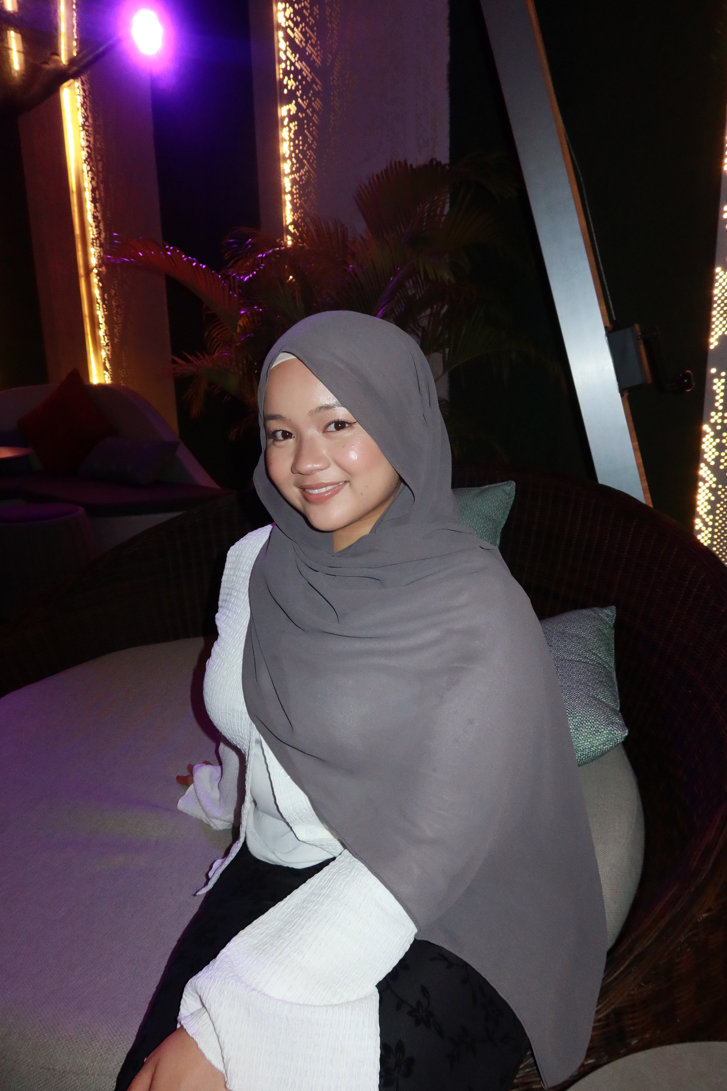 A smiling young woman wearing a grey hijab, white top, and black pants, sitting in a dimly lit lounge with colorful lights and a plant in the background.