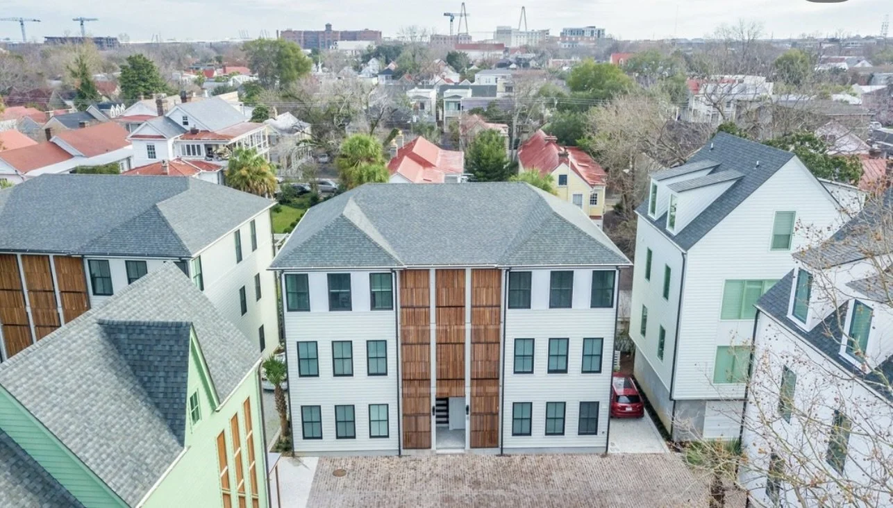 Aerial view of a neighborhood showing three multi-story houses with different exterior accents, surrounded by trees and other homes, with some cranes visible in the distance.