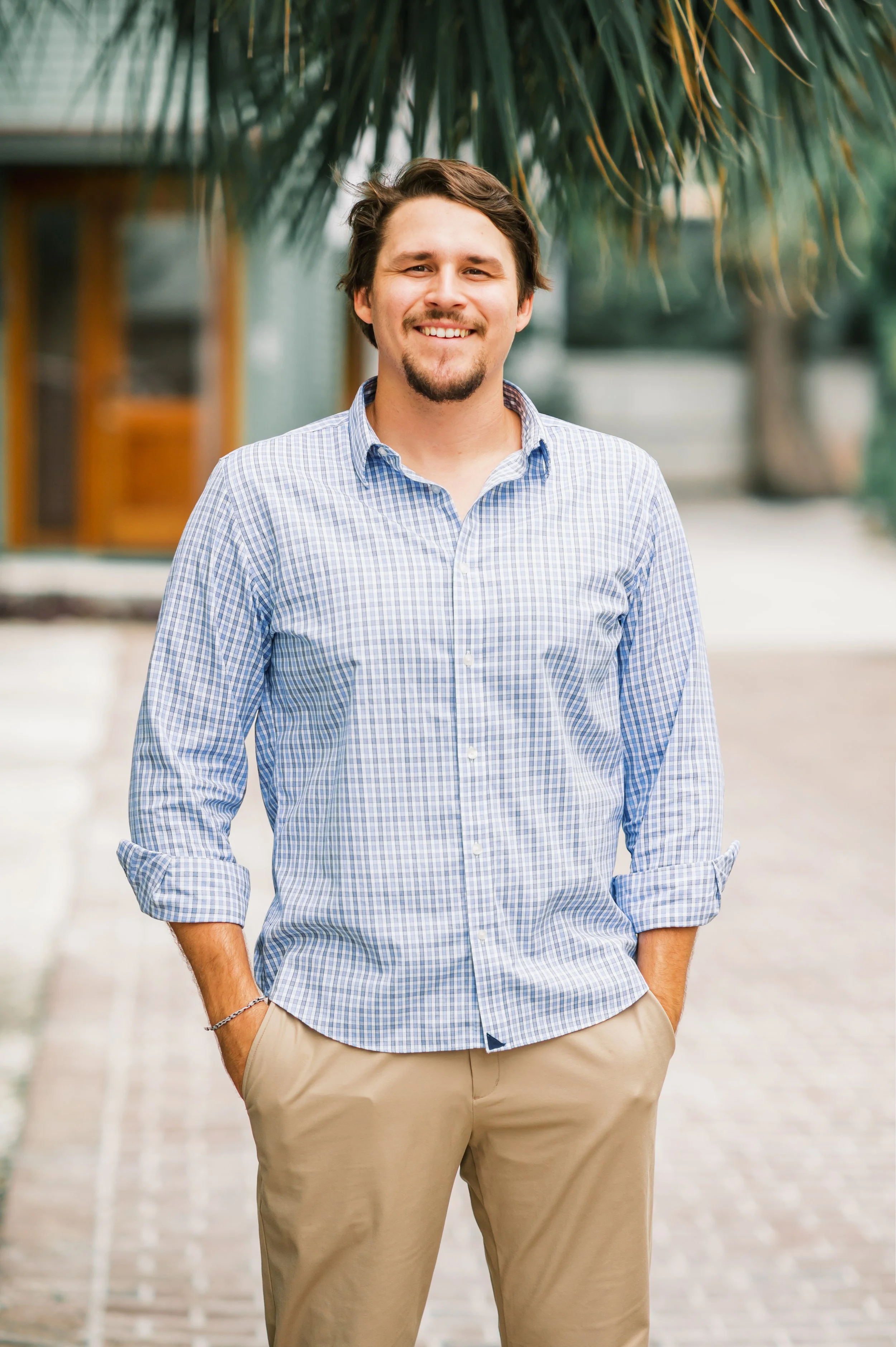 A man in a blue checkered shirt and khaki pants standing outdoors with a smile, with green palm leaves overhead and buildings in the blurred background.