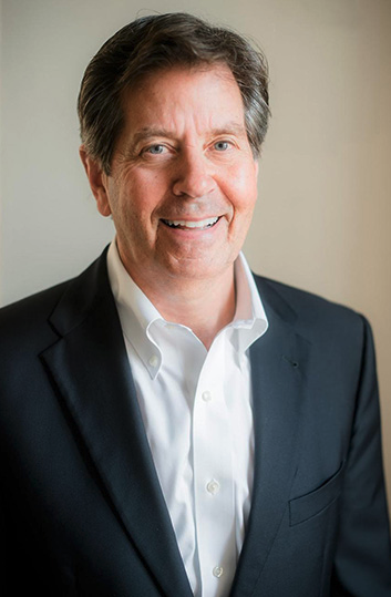 A smiling middle-aged man with brown hair and blue eyes, wearing a navy blazer and white shirt, posed against a neutral background.