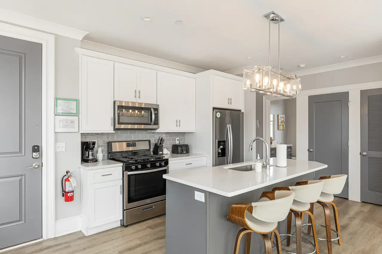 Modern kitchen with white cabinets, gray walls, stainless steel appliances, marble countertop island with four barstools, hanging light fixture, and a door with a gray finish.