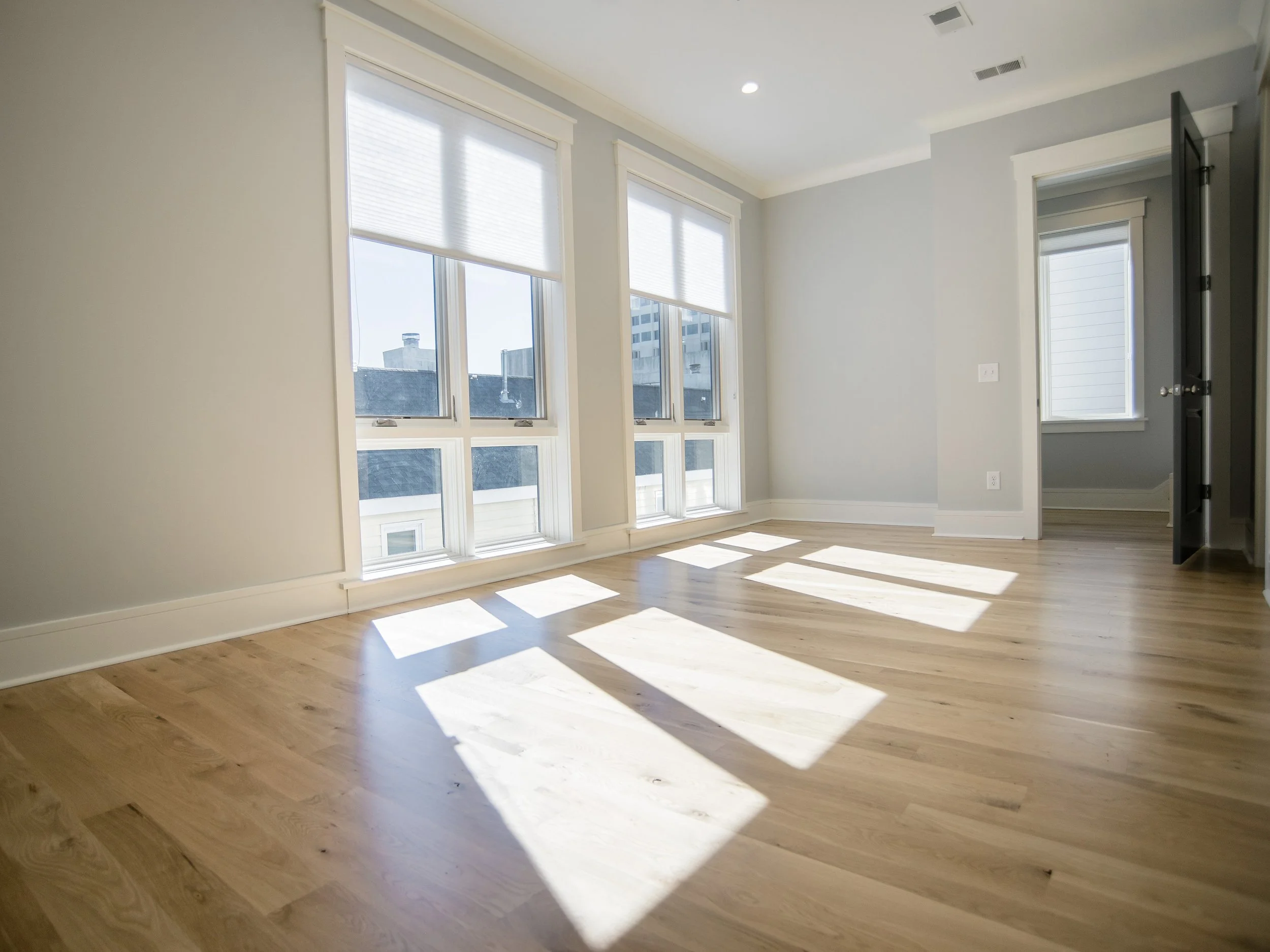 Empty room with large windows, light-colored walls, wooden floor, and sunlight casting shadows on the floor.