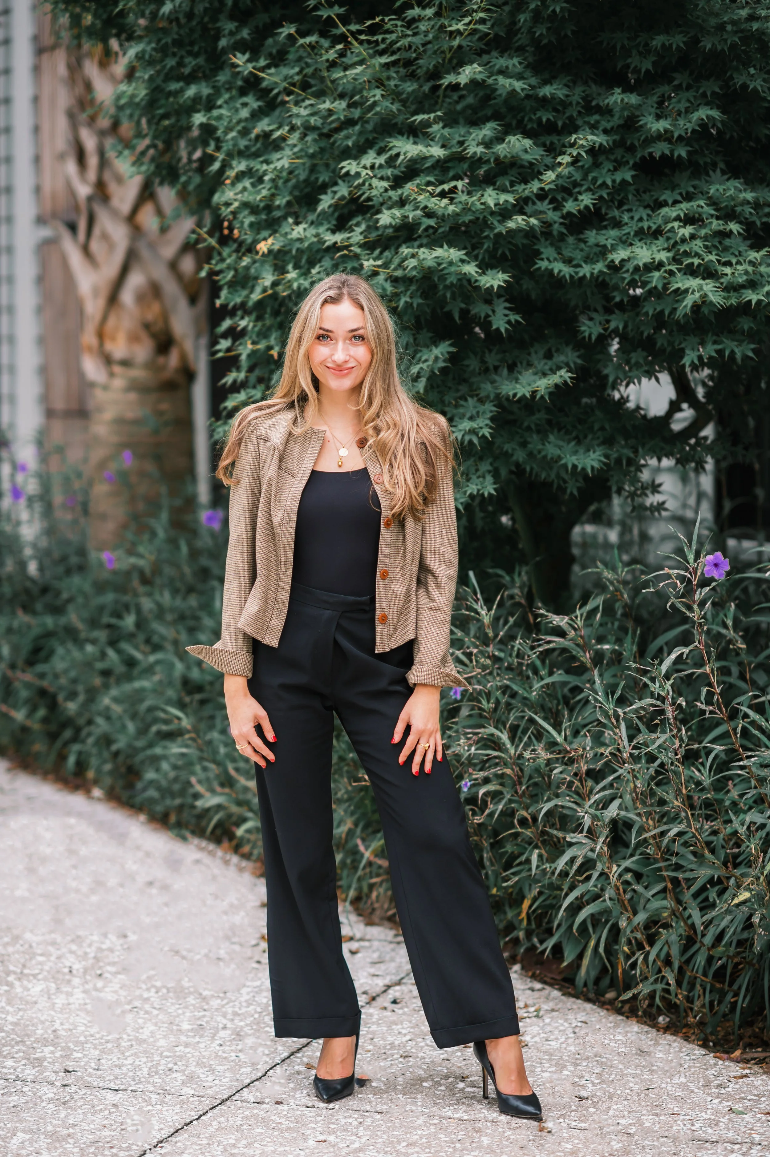 A woman standing outdoors on a concrete pathway near green leafy plants. She has long blonde hair, a beige blazer over a black top, black trousers, and black high heels. She is smiling at the camera.