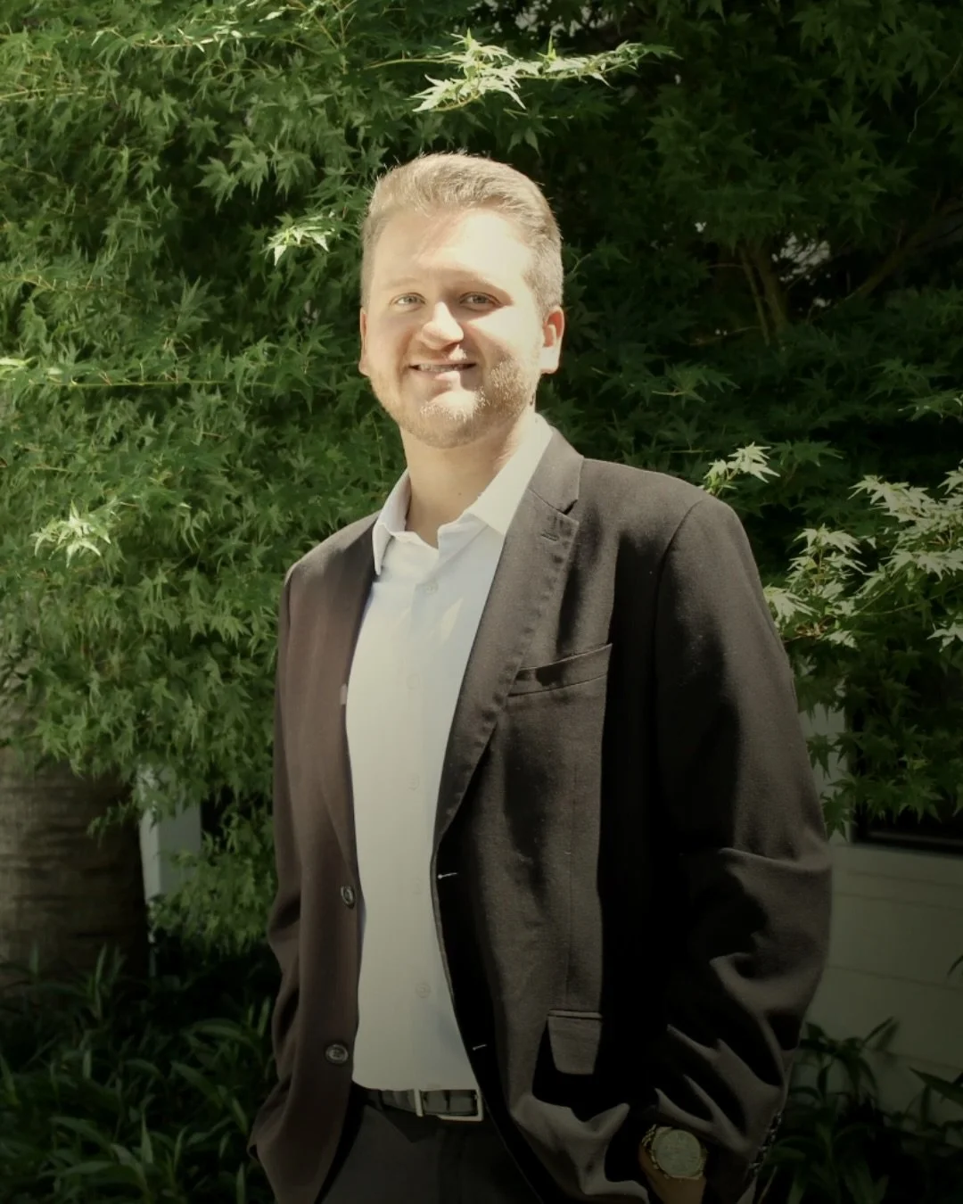 A young man with light hair and beard in a white dress shirt and black suit jacket standing outdoors in front of green leafy background, smiling at the camera.