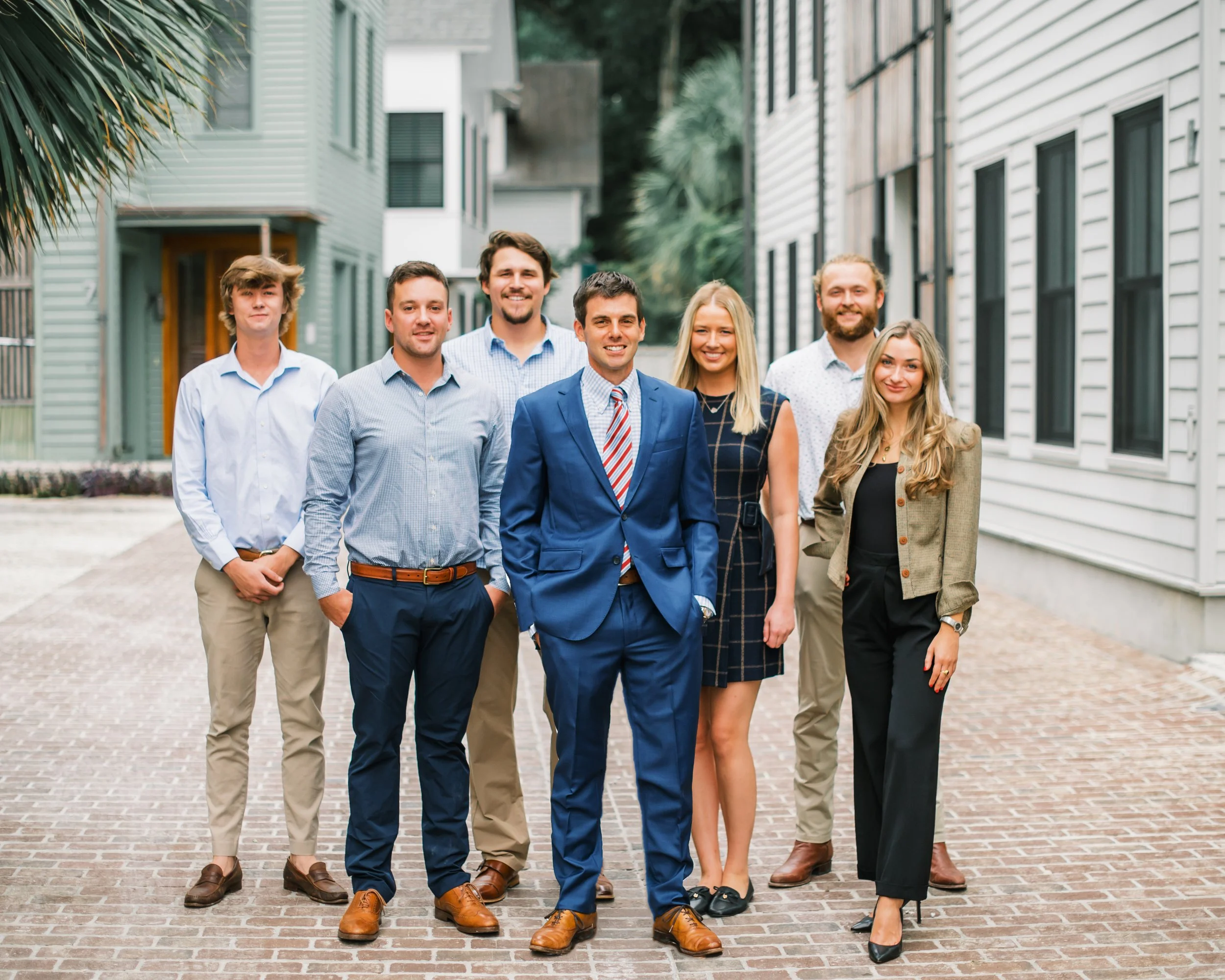 Group of seven diverse young professionals standing outdoors in front of modern buildings, smiling and looking at the camera.