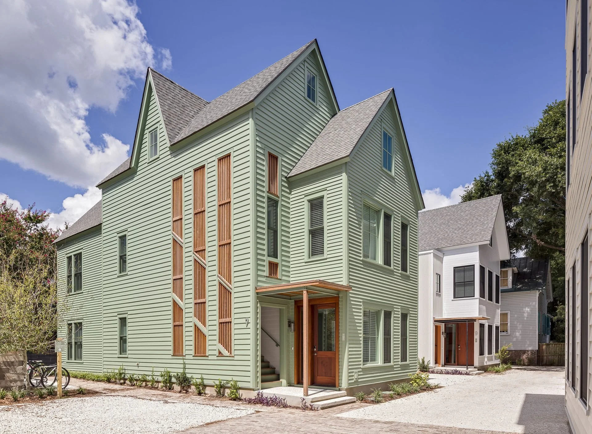 A modern green house with a steep gable roof, multiple windows, and wooden accents on the sides, situated in a neighborhood with similar style houses and a gravel pathway in front.