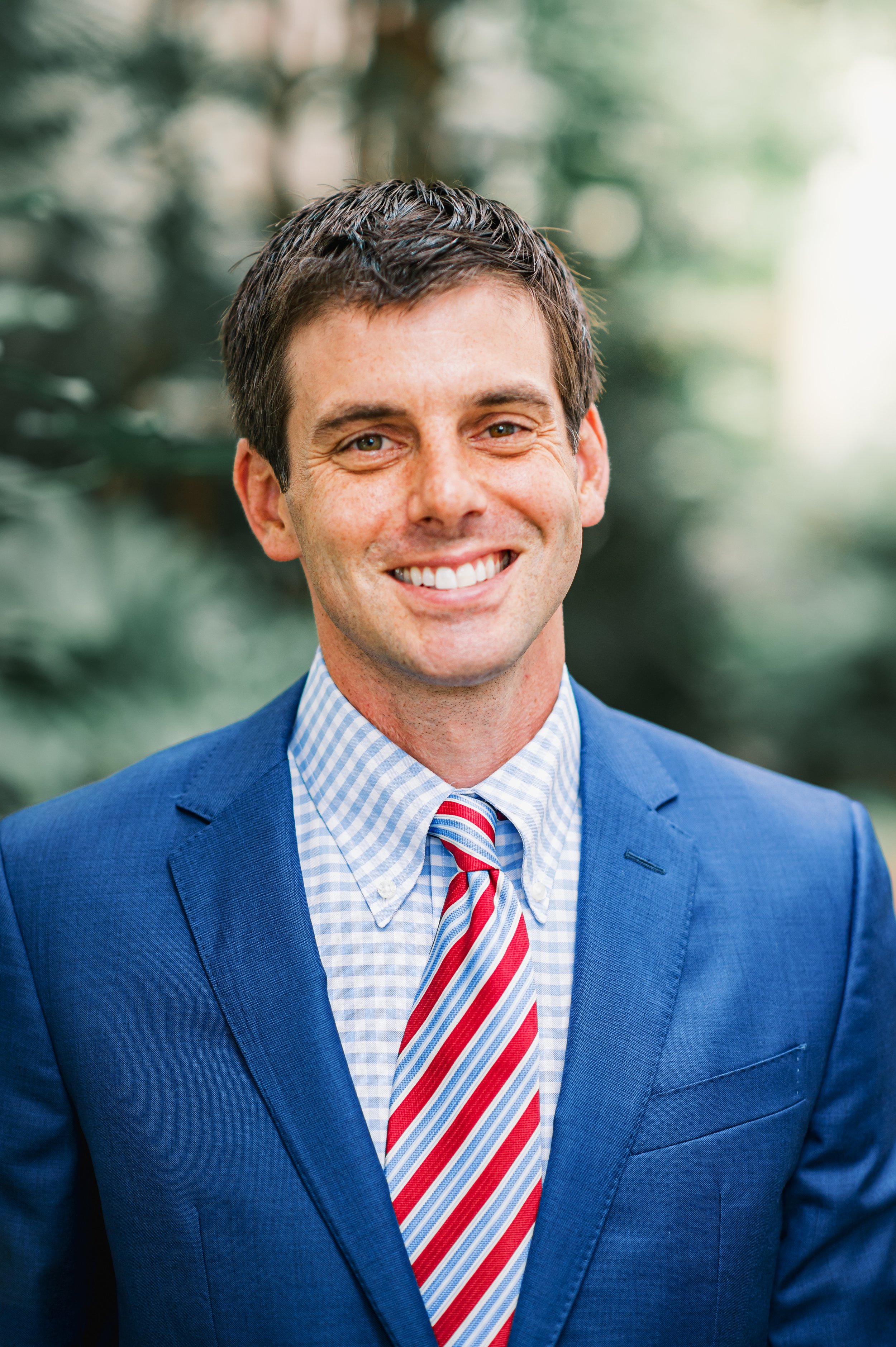 A smiling man in a blue suit, wearing a white and blue checkered shirt with a red and white striped tie, standing outdoors with blurred greenery in the background.