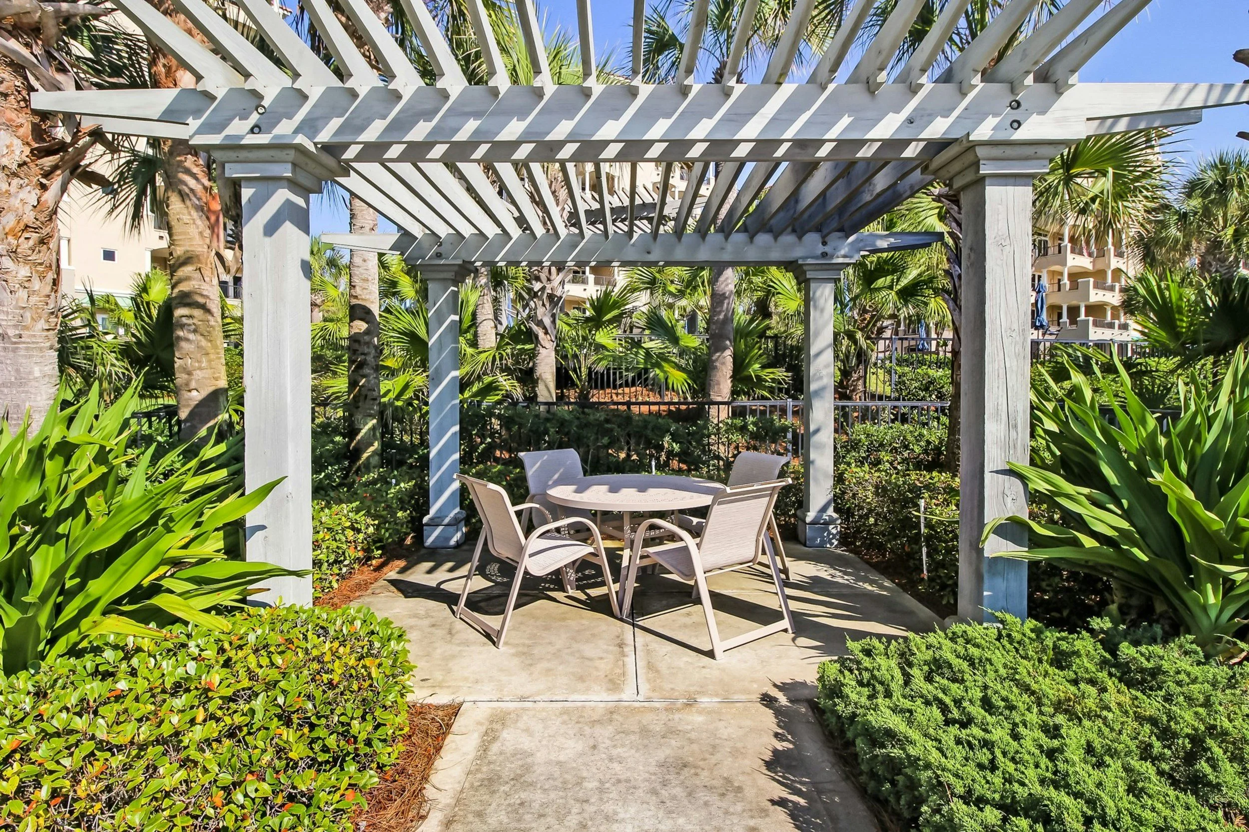 An outdoor patio area with a white wooden pergola, round table, and four beige chairs, surrounded by lush green plants and palm trees, with a building in the background.