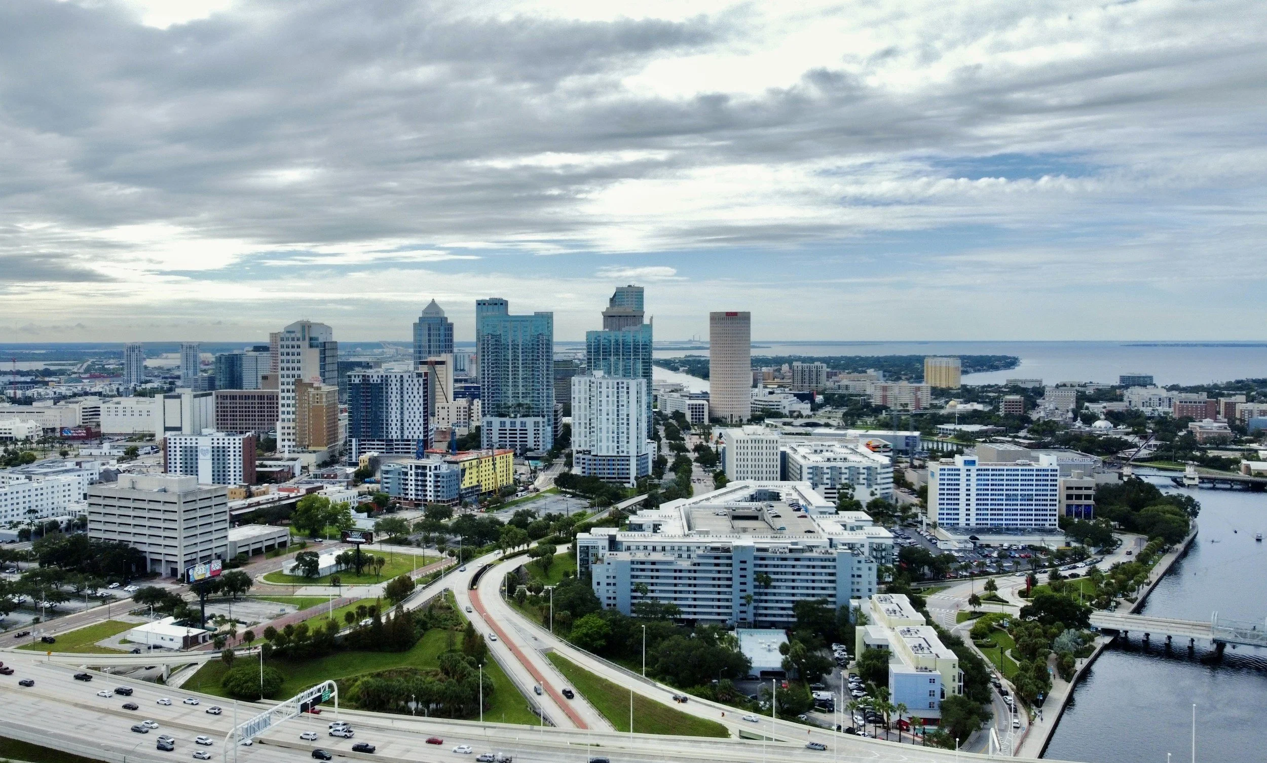 A city skyline with tall modern buildings, a body of water, and a cloudy sky above.