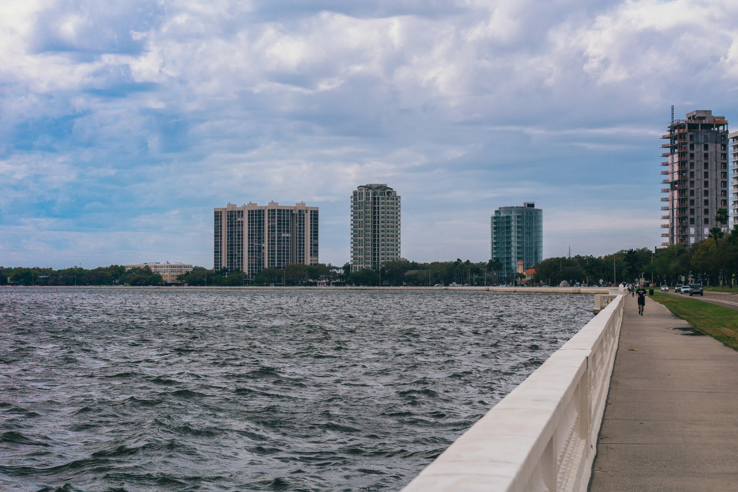 City skyline with tall buildings viewed across a body of water, with a sidewalk along the water's edge and a partly cloudy sky overhead.