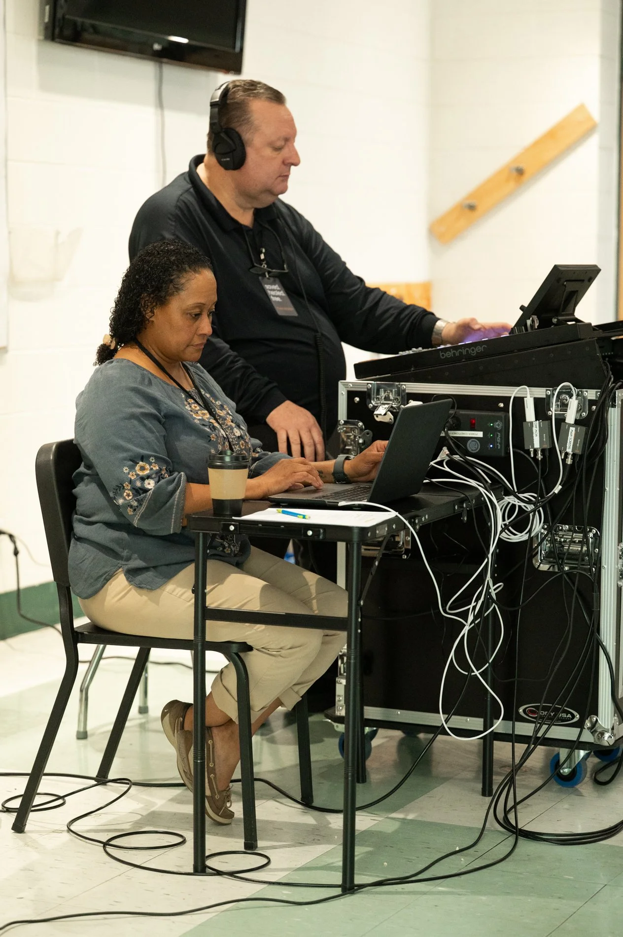 Two people working with audio and video equipment; a woman seated at a desk with a laptop and a coffee cup, and a man standing beside her with headphones and a keyboard.
