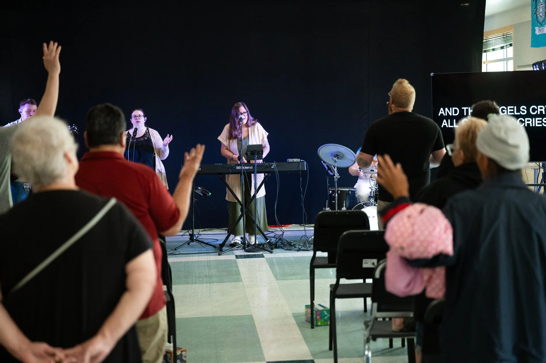 A group of people attending a church service or concert, listening to a live band perform on stage, with some raising their hands in worship or appreciation.