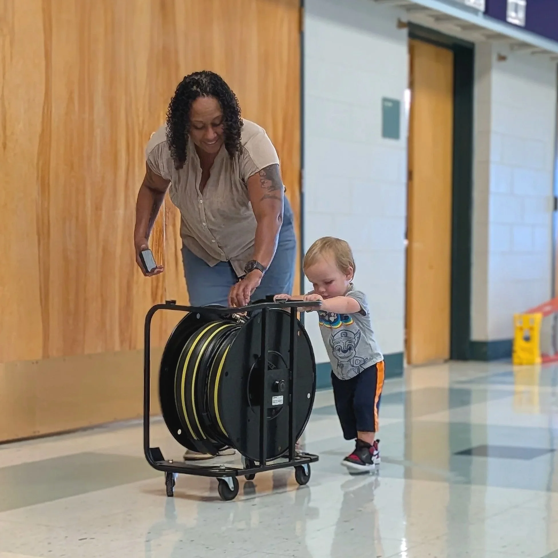 A woman with curly black hair and tattoos on her arm is smiling and bending over a luggage cart with wheeled suitcases, seemingly talking to a young boy with blond hair and a patterned shirt, in a hallway with light-colored tile floors and wooden wall paneling.