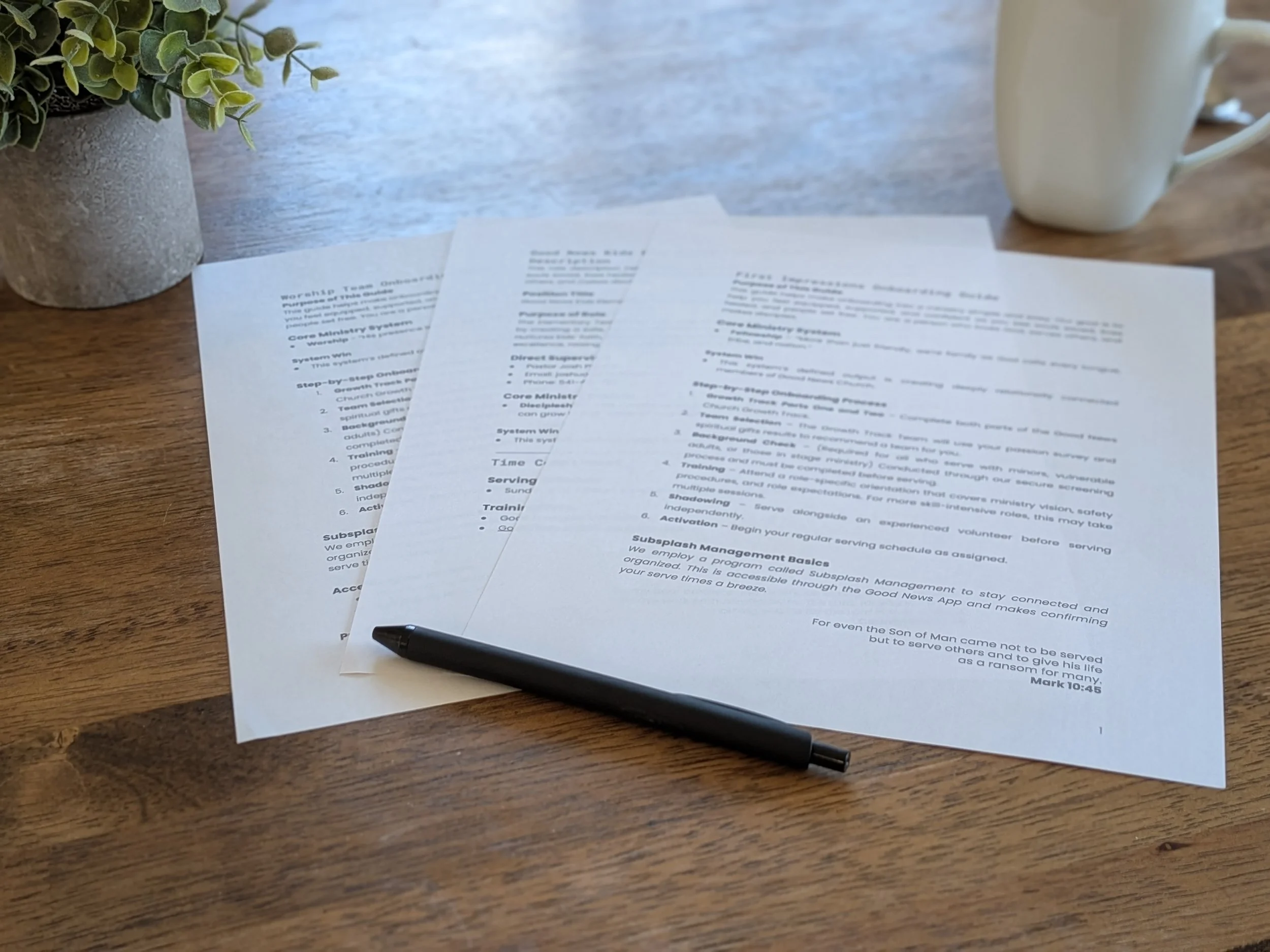 A wooden table with three sheets of paper containing text, a black pen resting on the lower right sheet, a potted green plant in a textured gray pot on the left, and a white mug in the background on the right.