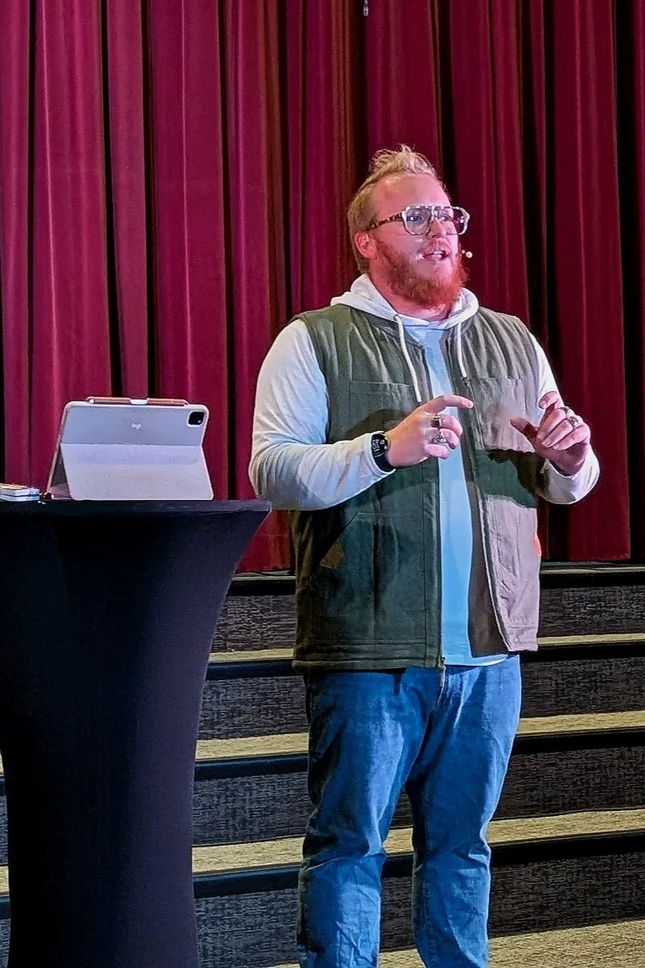 A man with glasses, a beard, and reddish hair giving a presentation on stage with a red curtain backdrop, wearing a hoodie, vest, and jeans, standing beside a table with a tablet.