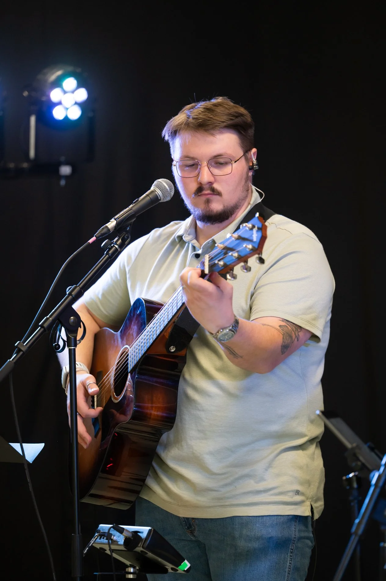 A man with glasses and tattoos on his left arm is playing an acoustic guitar and singing into a microphone. He is standing in a dark room with stage lighting in the background.
