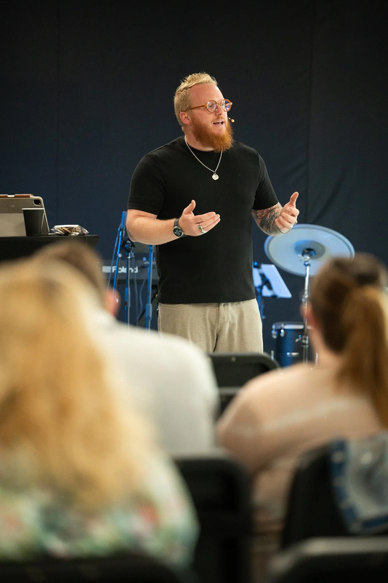 A man with blond hair, glasses, and a red beard speaking to an audience in a room equipped with musical instruments, including drums and a keyboard.