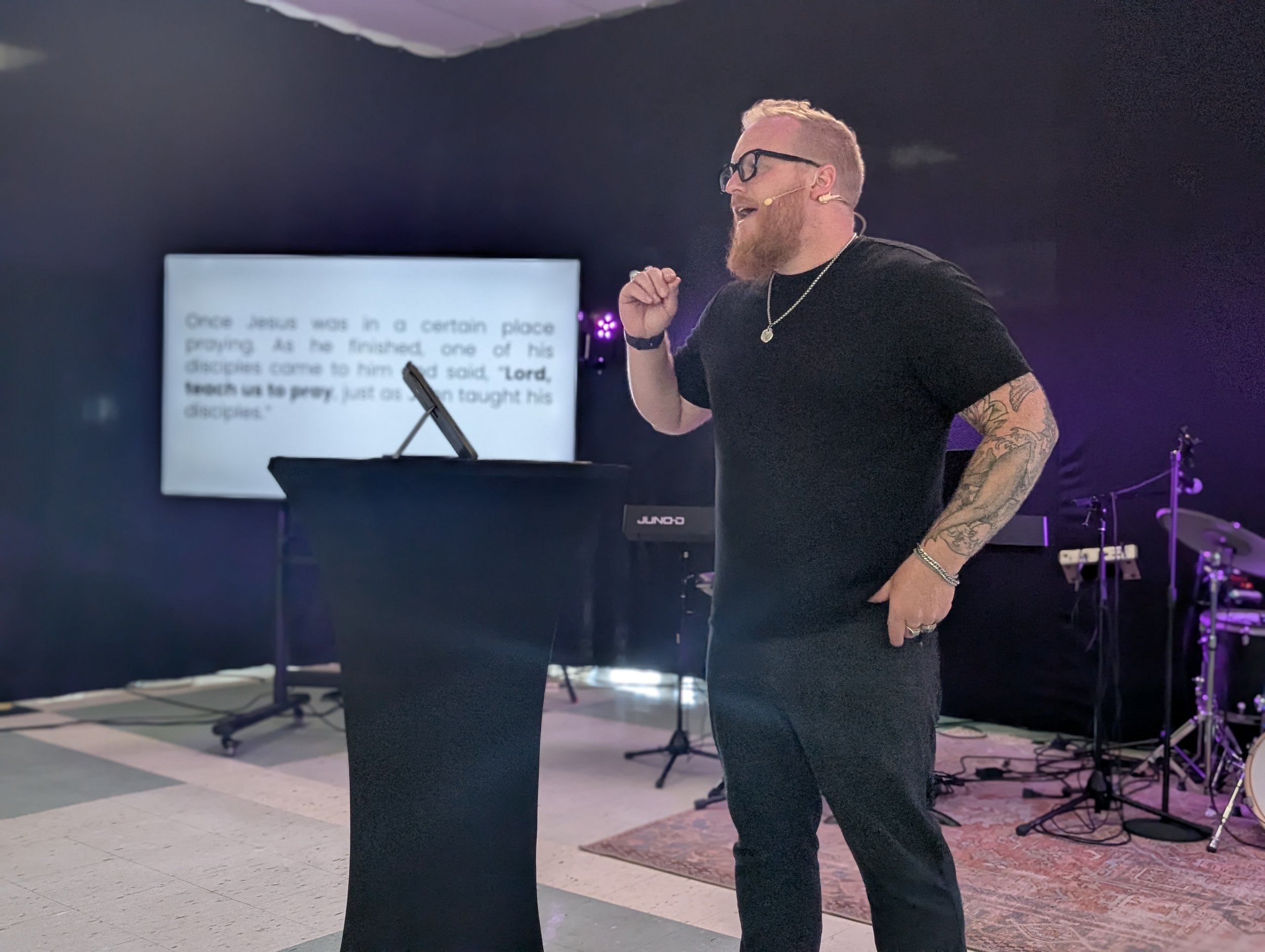 A man with light skin, glasses, a beard, and tattoos on his arm is speaking into a headset microphone. He is smiling and gesturing with his right fist. Behind him is a screen with blurred text and a music stand, with musical instruments nearby, indicating a stage or presentation setting.