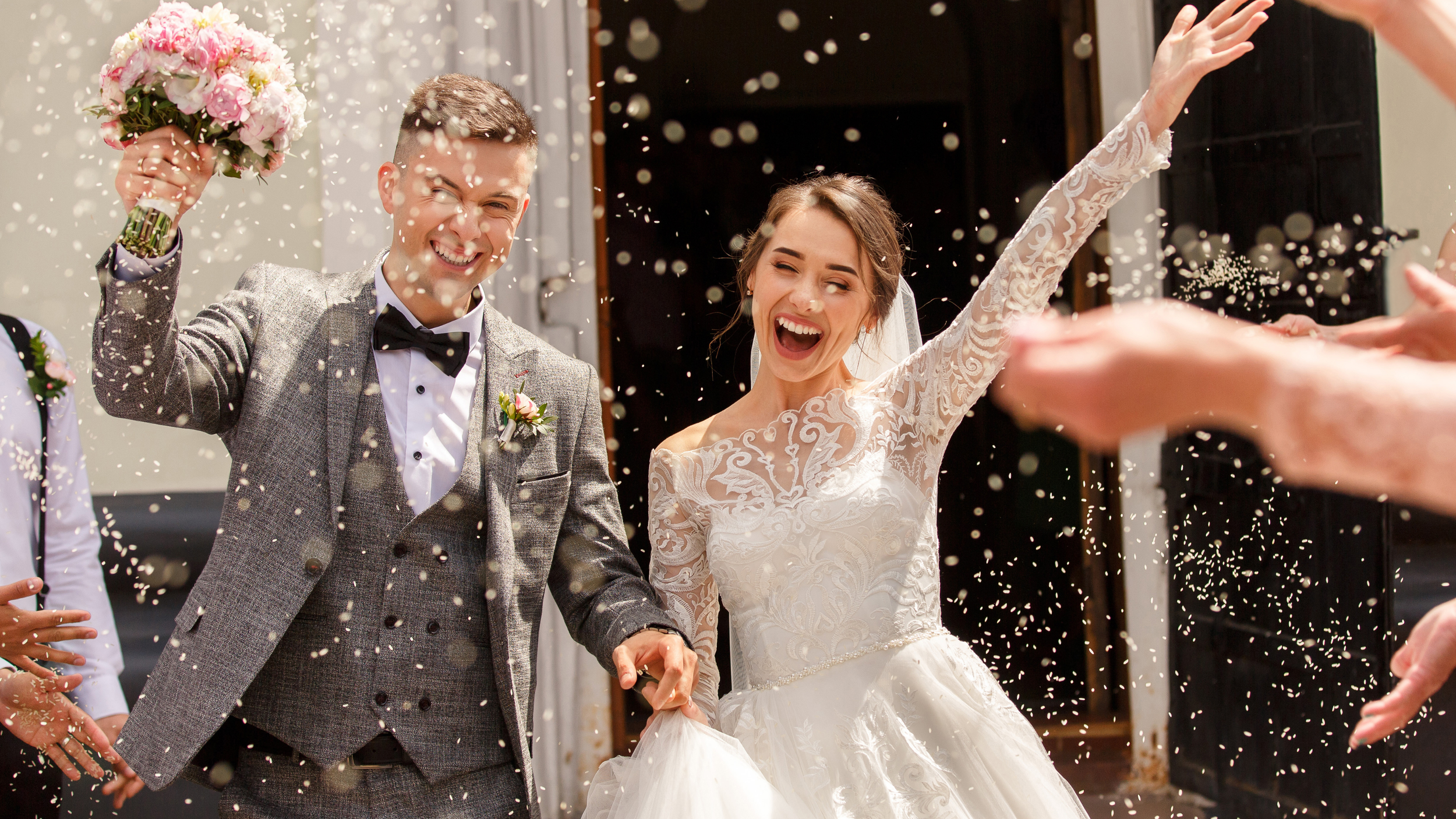 Ein frisch verheiratetes Paar, ein Mann in Anzug und eine Frau in einem weißen Hochzeitskleid und traumhaften Eheringen, wird durch Konfetti begrüßt, während sie lächelnd und fröhlich sind.