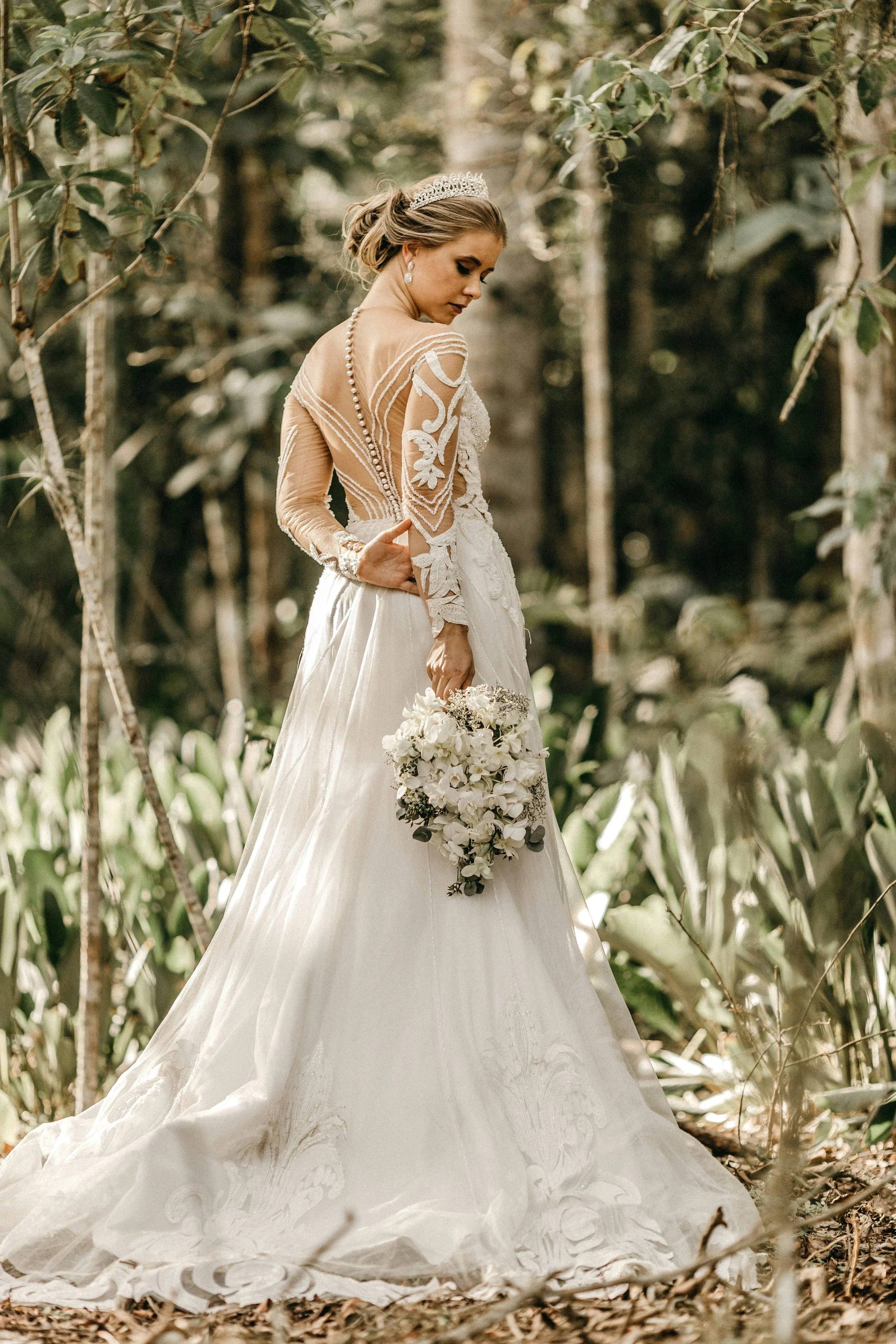 Eine Frau in einem weißen Brautkleid mit Stickereien und Spitze, trägt besonderen Brautschmuck, darunter Perlenohringe, Haarschmuck, Brillanten und ein Armband