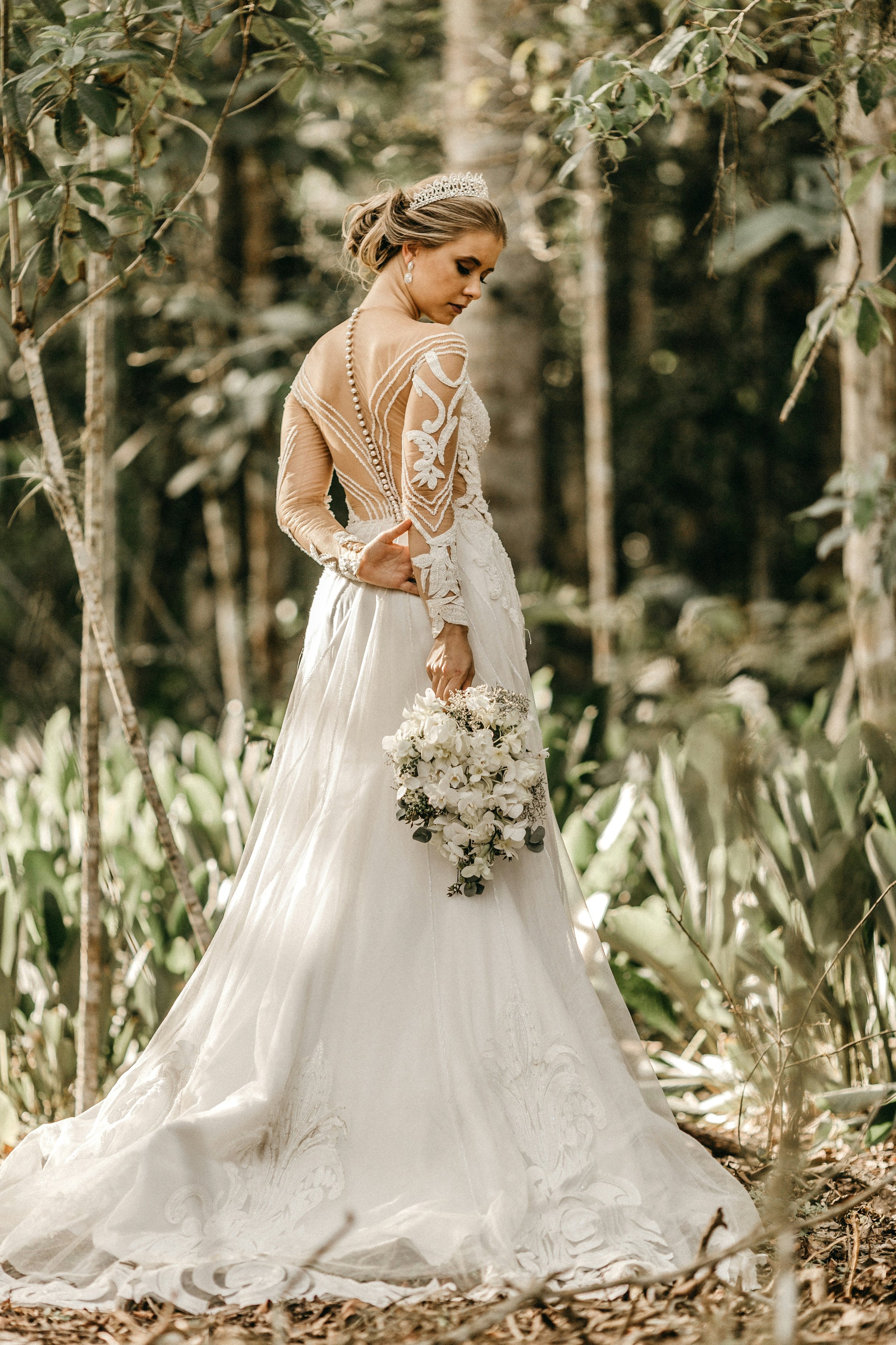 Eine Frau in einem weißen Brautkleid mit Stickereien und Spitze, trägt besonderen Brautschmuck, darunter Perlenohringe, Haarschmuck, Brillanten und ein Armband